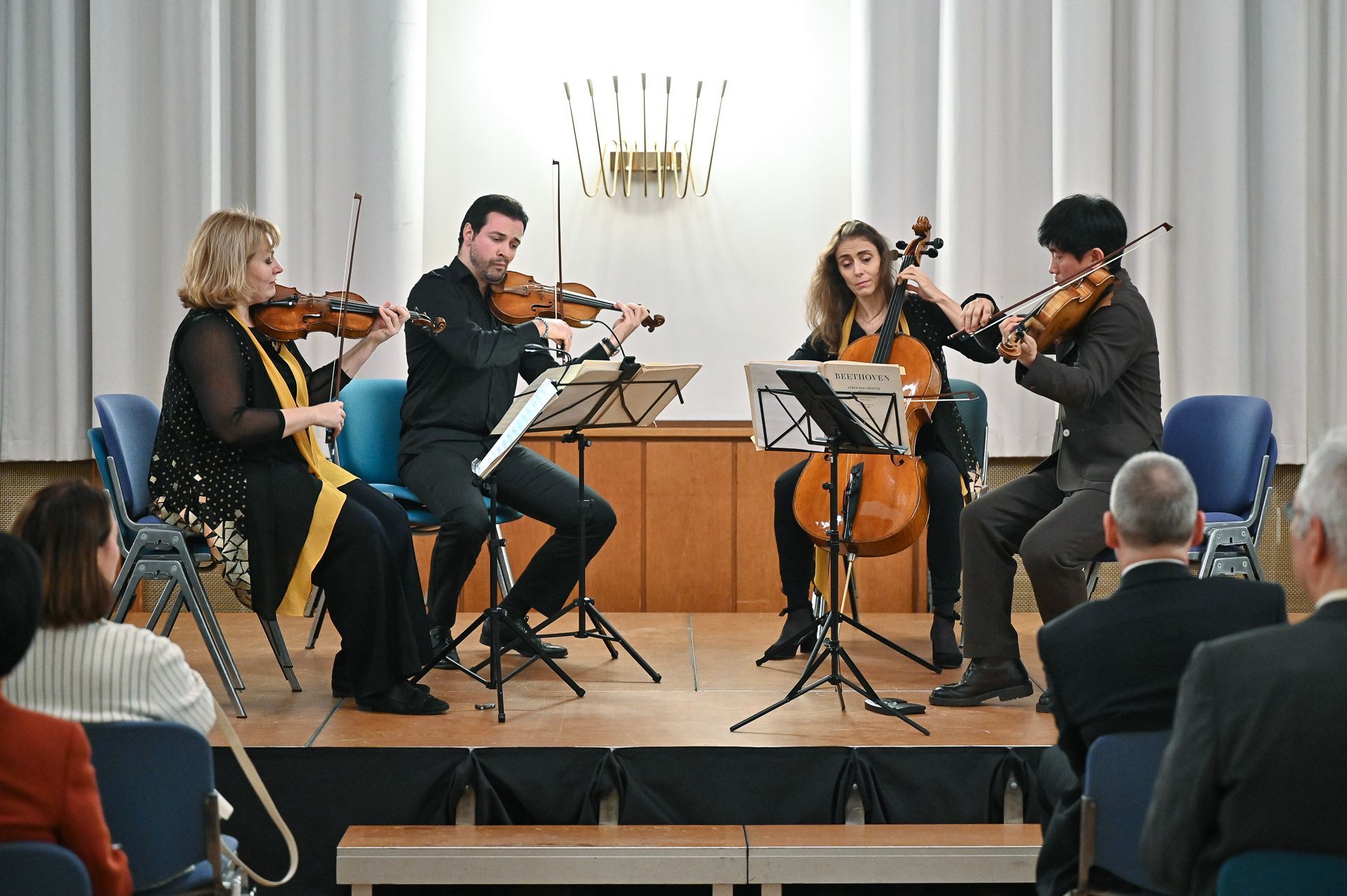Ein Streichquartett spielt auf der Bühne; zwei Violinisten, ein Cellist und eine Violinistin spielen ihre Instrumente. Das Publikum sitzt.