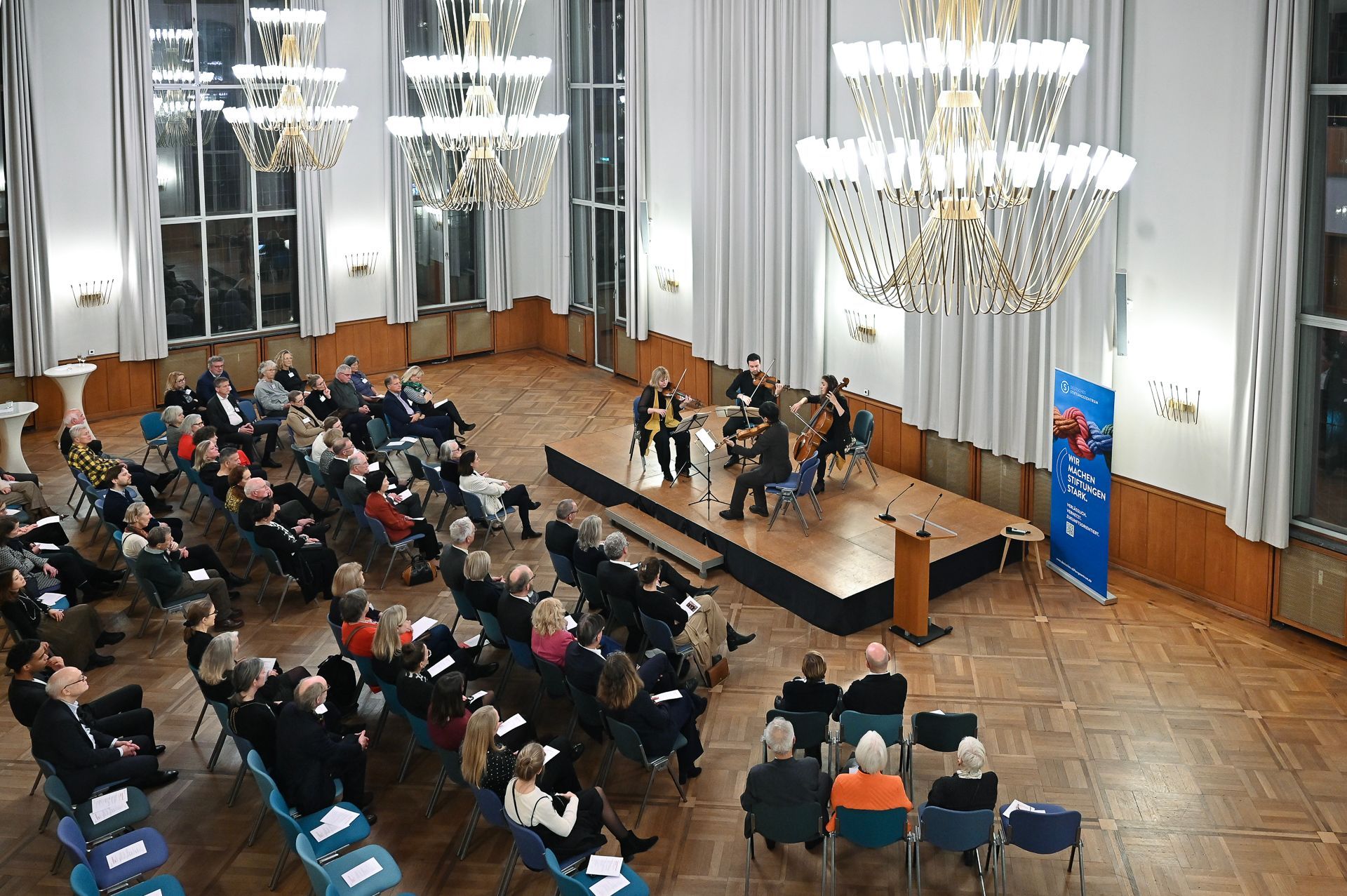 Publikum bei einer formellen Veranstaltung; Musiker auf der Bühne unter Kronleuchtern.