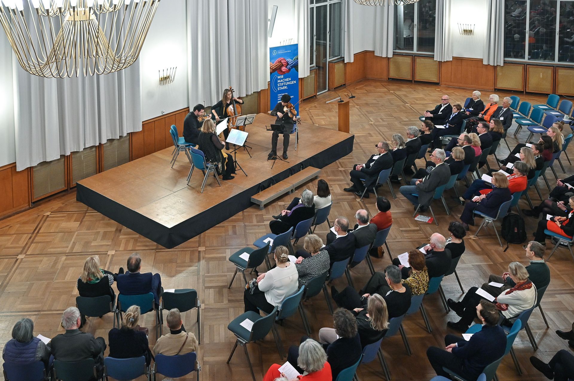 Podiumsdiskussion auf der Bühne, Publikum sitzt. Heller Raum, Bühne mit Rednern, Zuhörer.