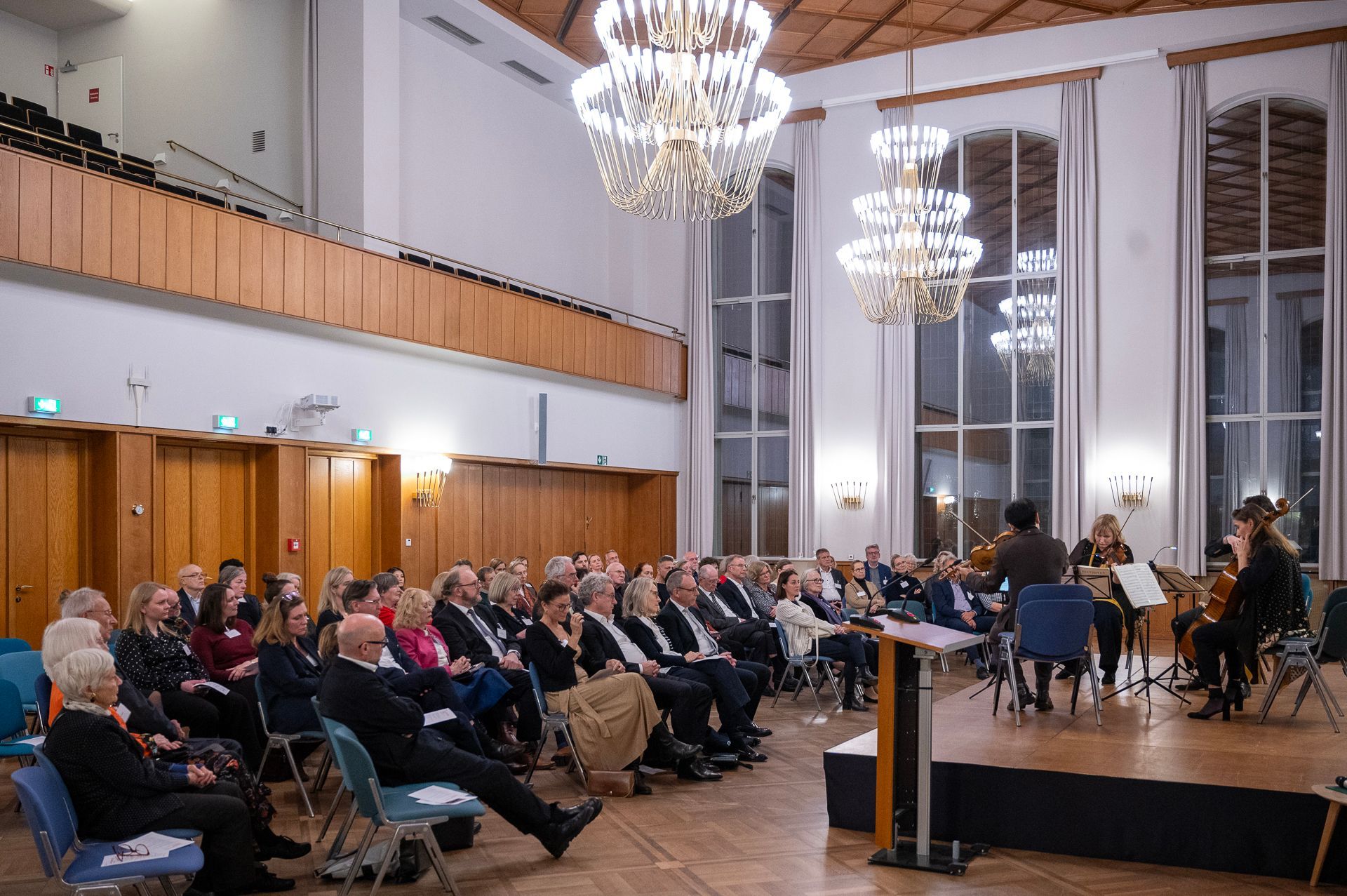 Zuschauer beobachten ein Streichquartett, das in einem großen Saal mit Kronleuchtern auf der Bühne auftritt.