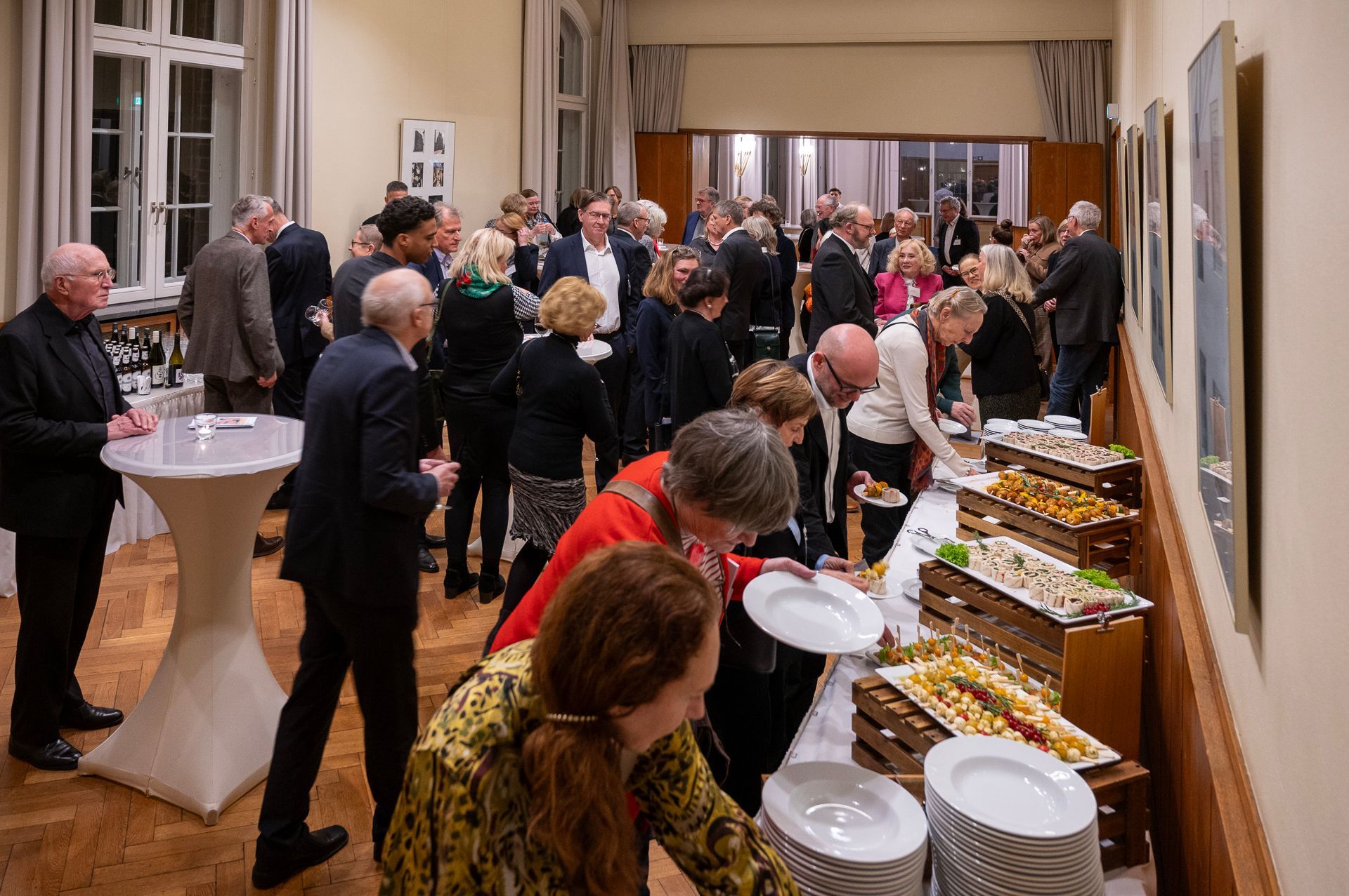 Menschen an einem Buffet in einem warm beleuchteten Raum. Gäste unterhalten sich angeregt an einem Tisch mit Speisen.