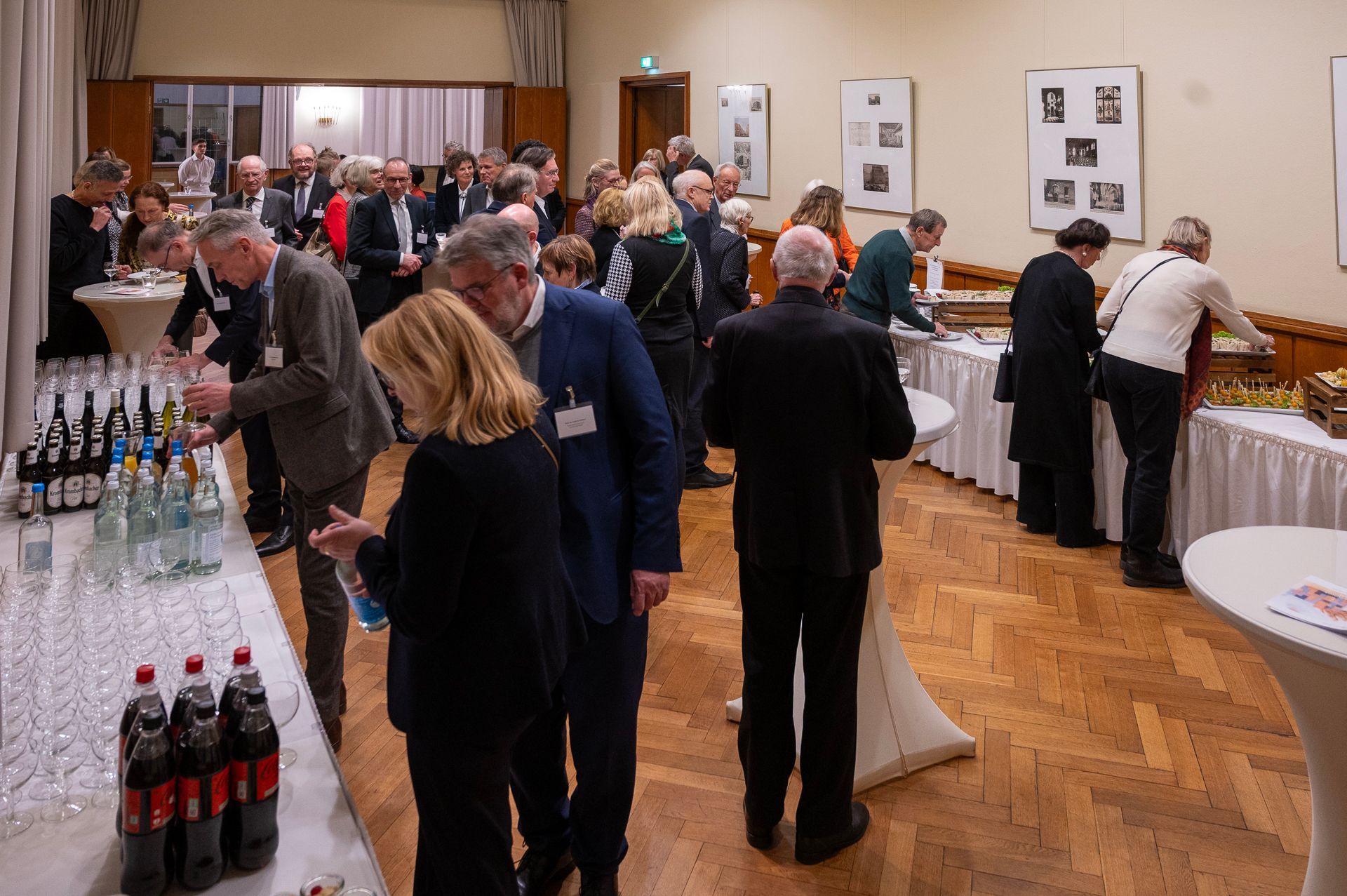 Menschen bei einem Empfang mit Getränken und Snacks in einem Saal mit Holzboden. Einige unterhalten sich, andere holen sich Getränke.