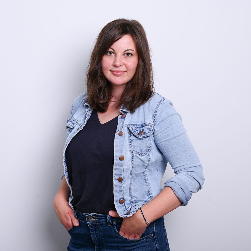 Frau in Jeansjacke, Hände in den Taschen, lächelt.