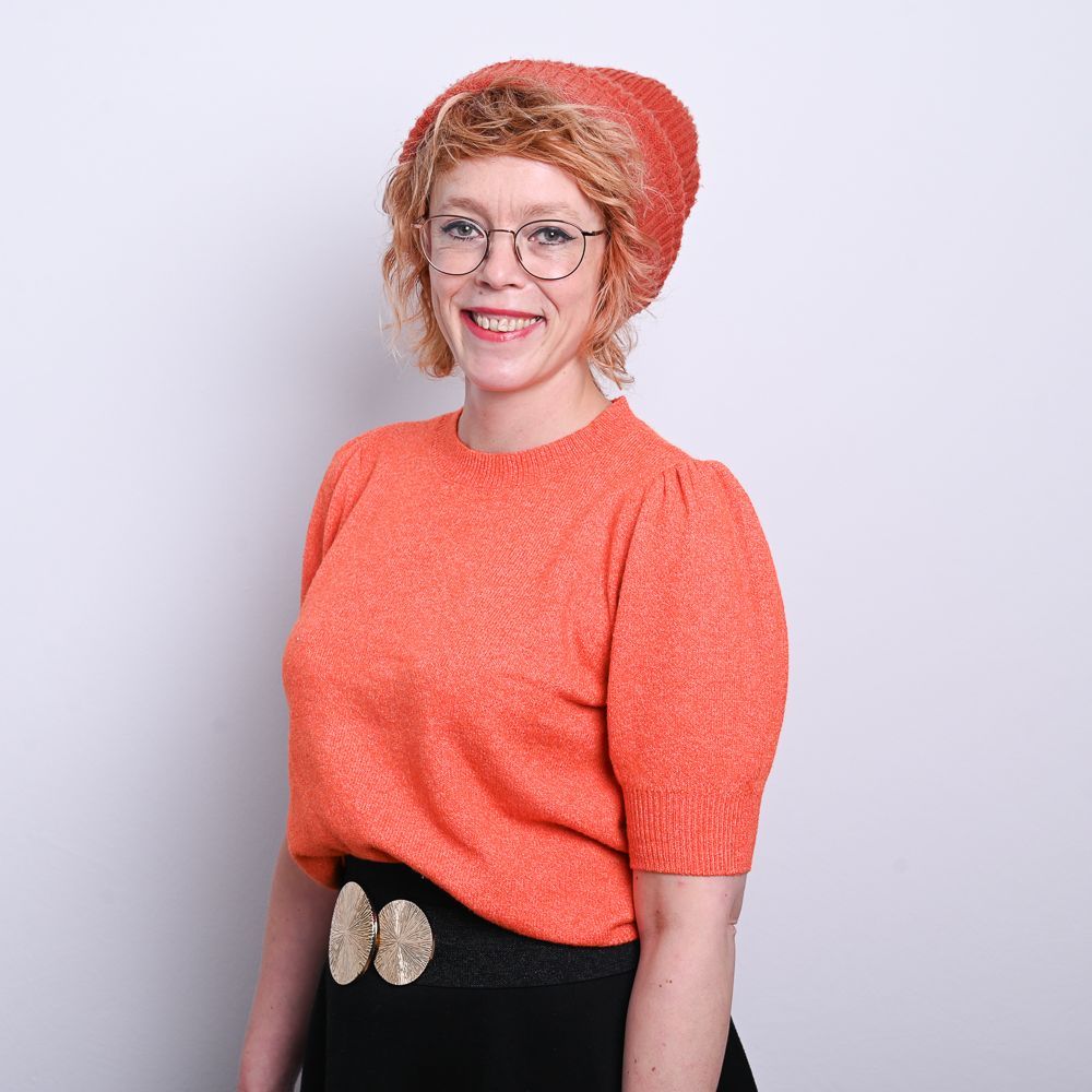 Frau in orangefarbenem Pullover und Hut, lächelnd, mit Brille.
