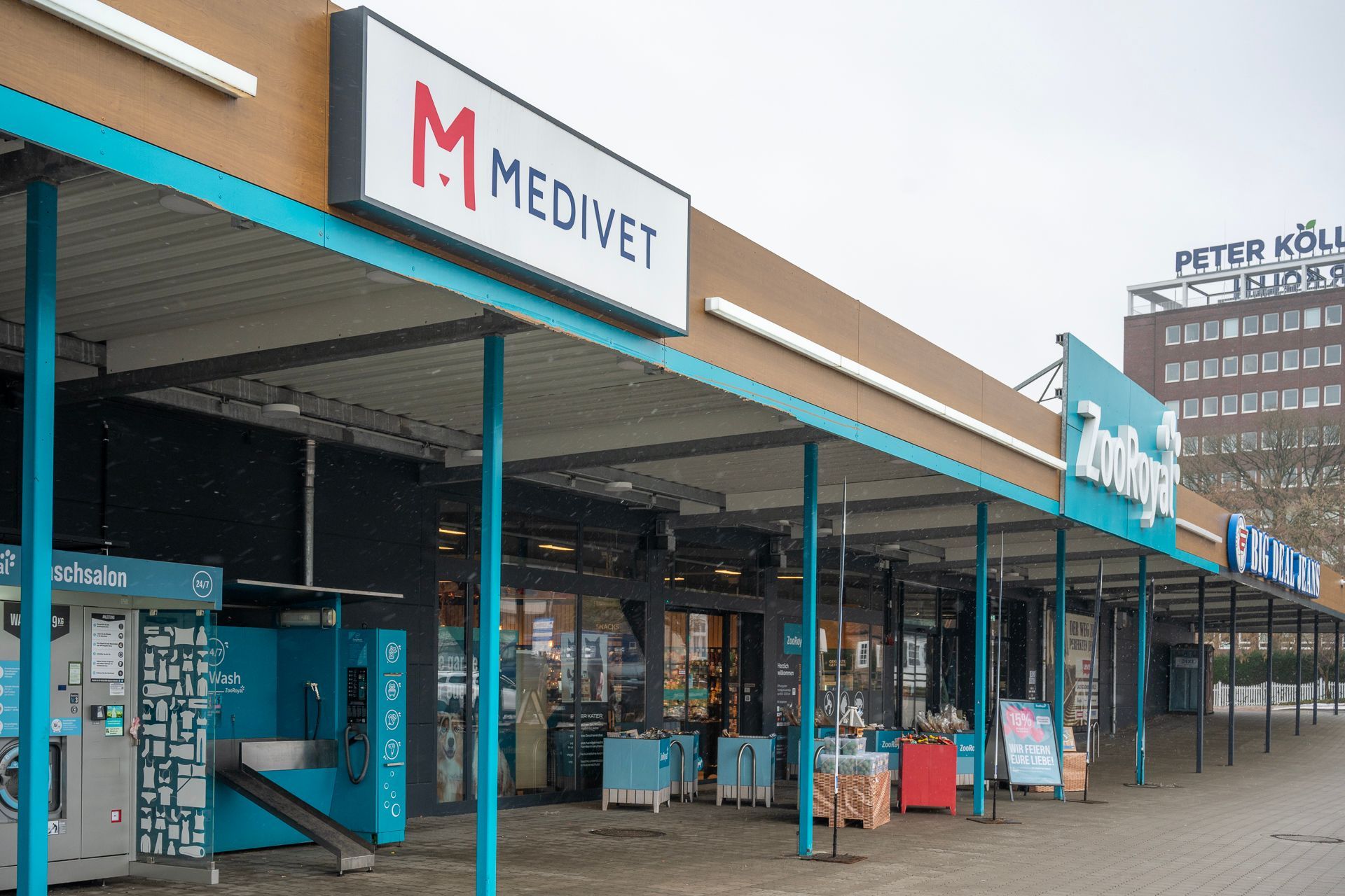 Eine Reihe von Geschäften unter einer türkisfarbenen Markise, darunter eine Medivet-Klinik, an einem bewölkten Tag.