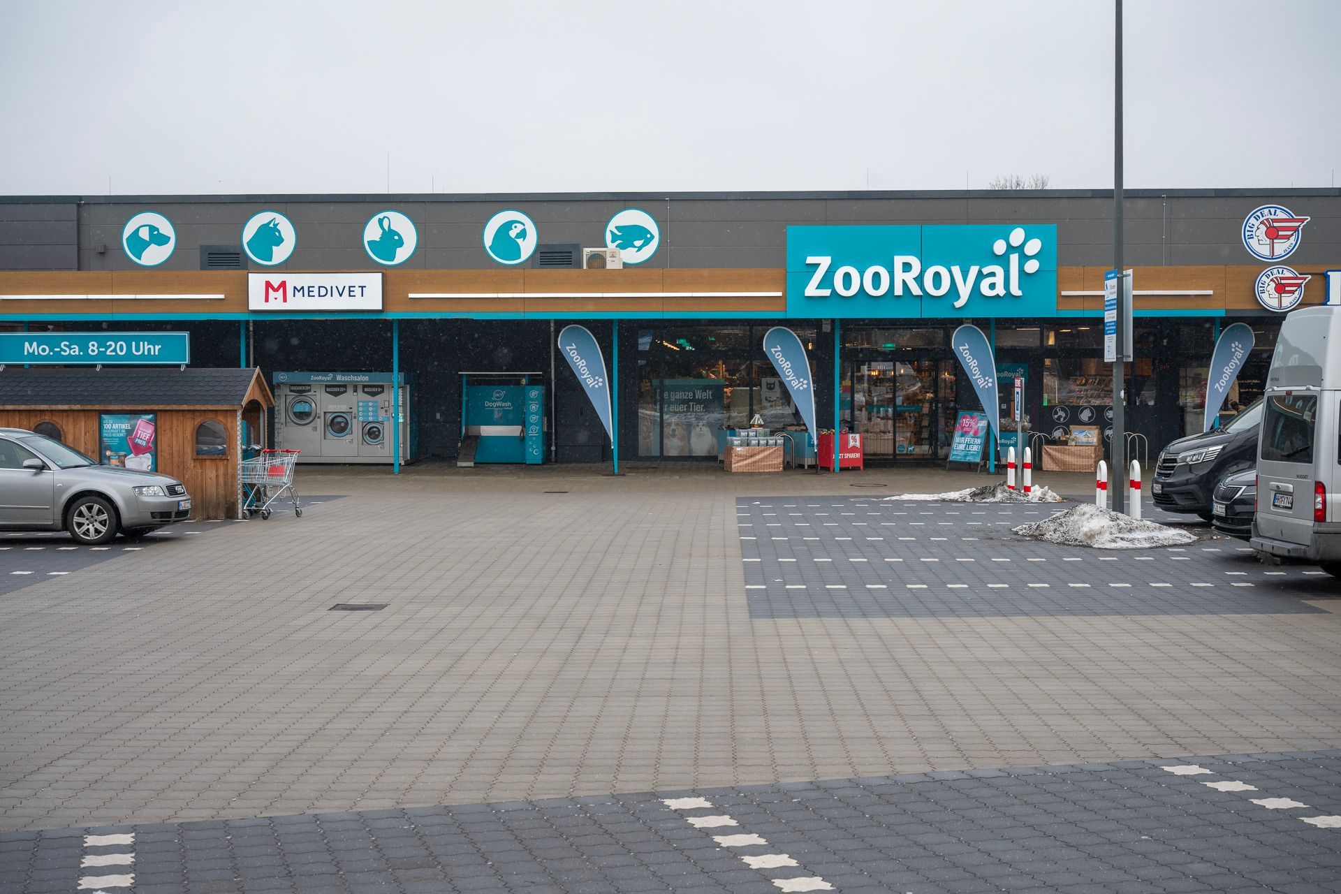 Außenansicht eines ZooRoyal-Tierbedarfsgeschäfts mit blauen Akzenten, Flaggen und davor geparkten Autos.