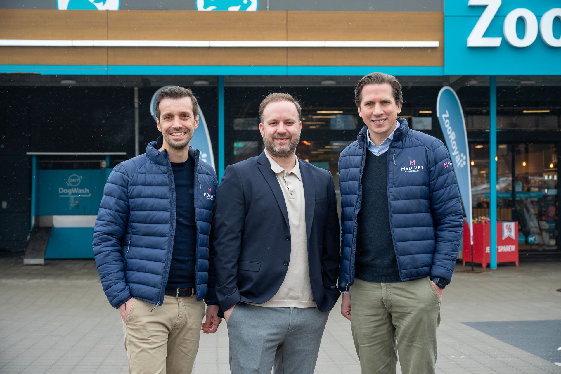 Drei Männer vor einem Zoo-Laden, zwei in blauen Jacken, einer in einem Blazer, alle lächelnd.