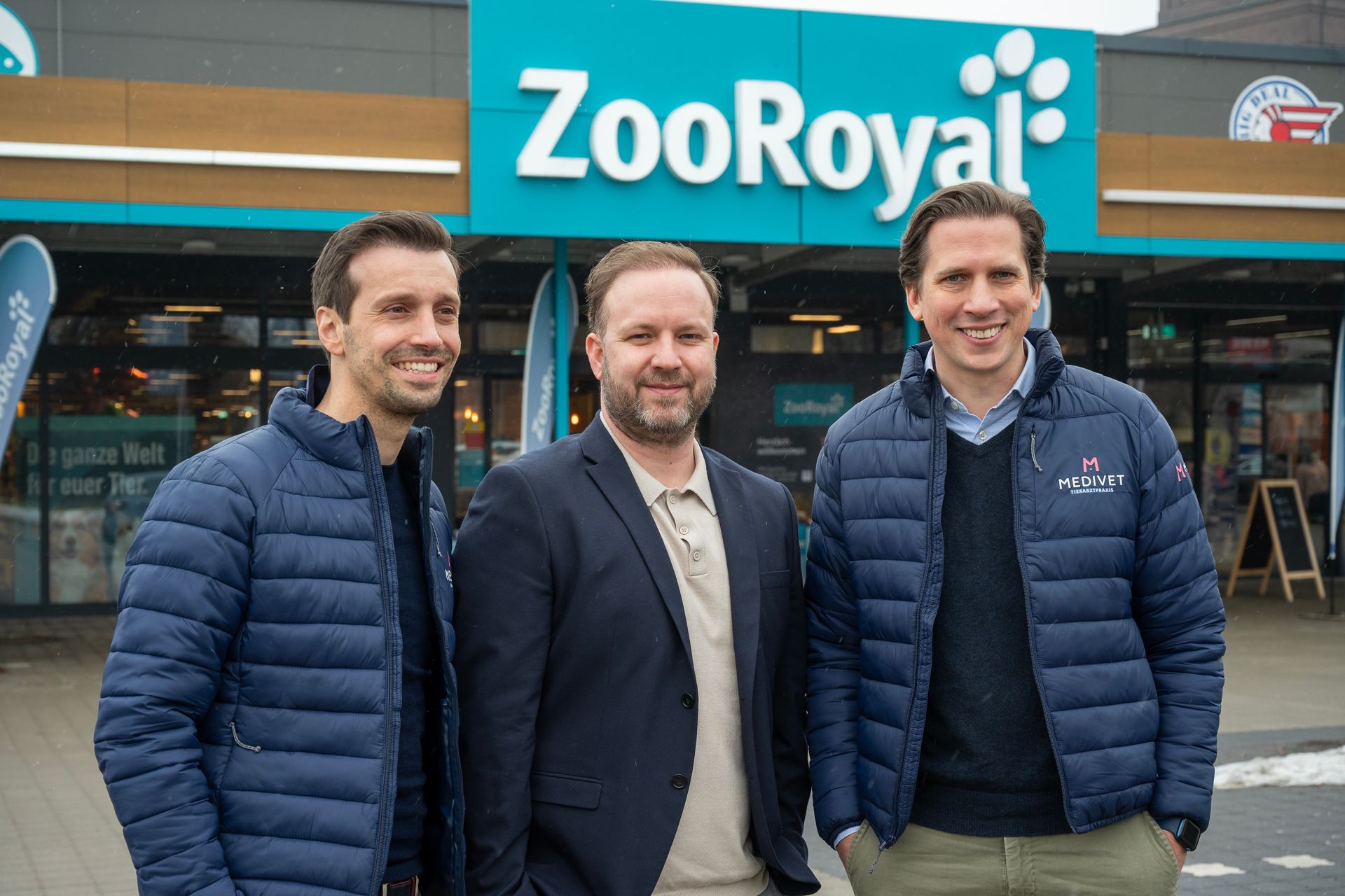 Drei Männer stehen lächelnd vor einem ZooRoyal-Laden. Blaue Jacken, im Freien.