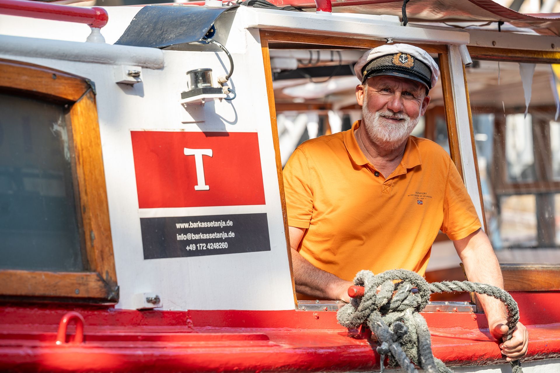 Ein Mann mit Kapitänsmütze lehnt lächelnd aus dem Fenster eines rot-weißen Bootes. Er trägt ein orangefarbenes Hemd, ein Seil ist sichtbar. Ein „T“ ist an der Bootsseite angebracht.