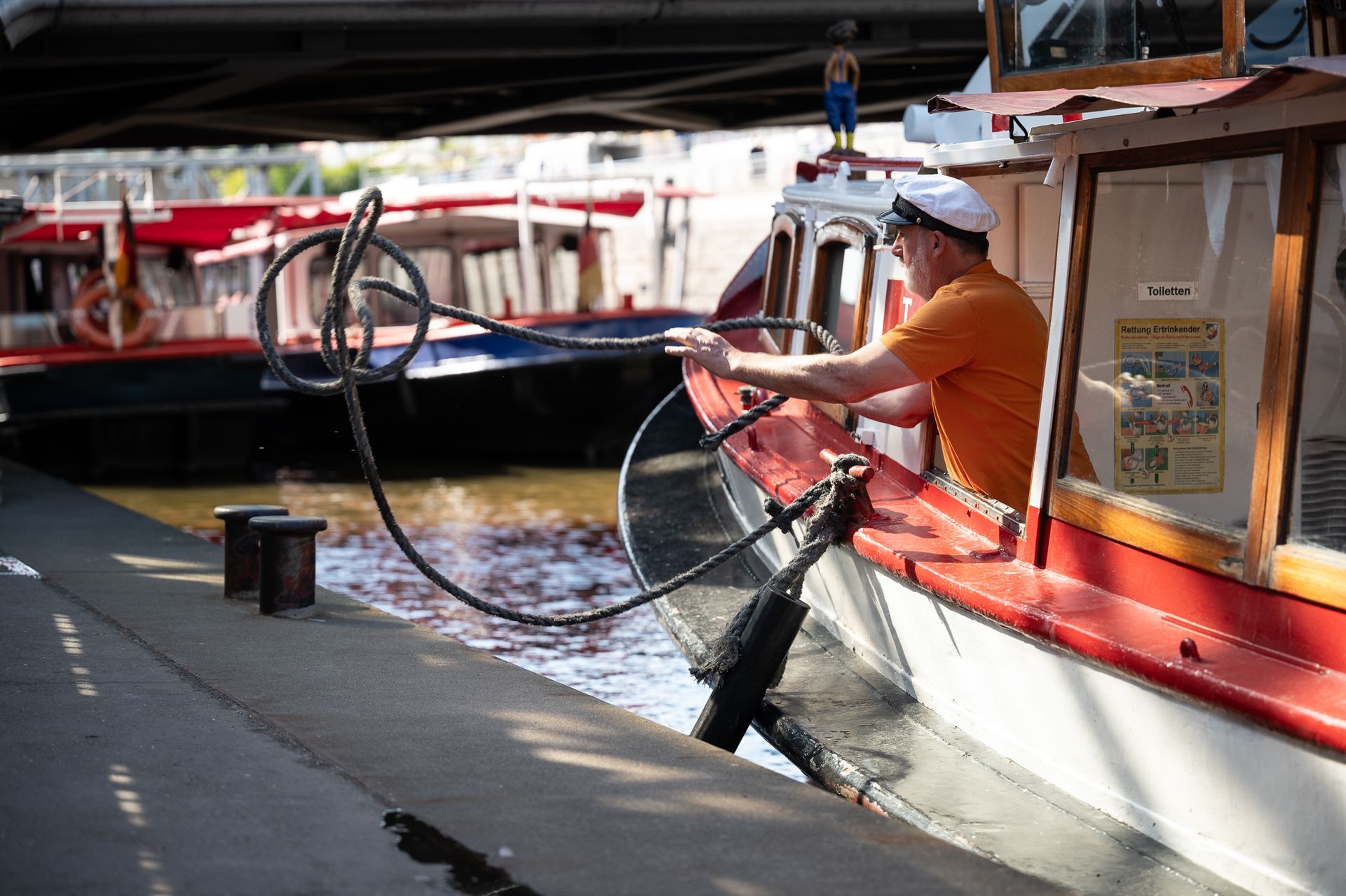 Ein Mann befestigt in einem Boot ein Seil an einem Dock; im Hintergrund sind weitere Boote zu sehen.