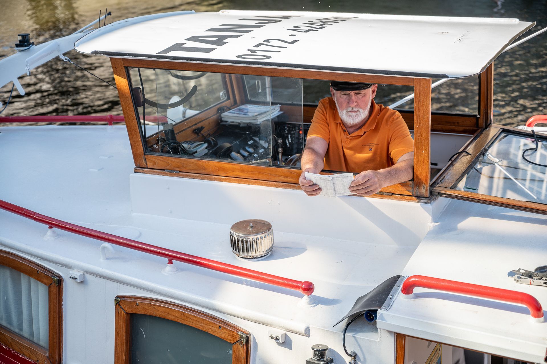 Ein Mann in orangefarbenem Hemd sitzt auf einem Boot und hält Geld. Das Boot hat ein Fenster, ein weißes Dach und rote Akzente.