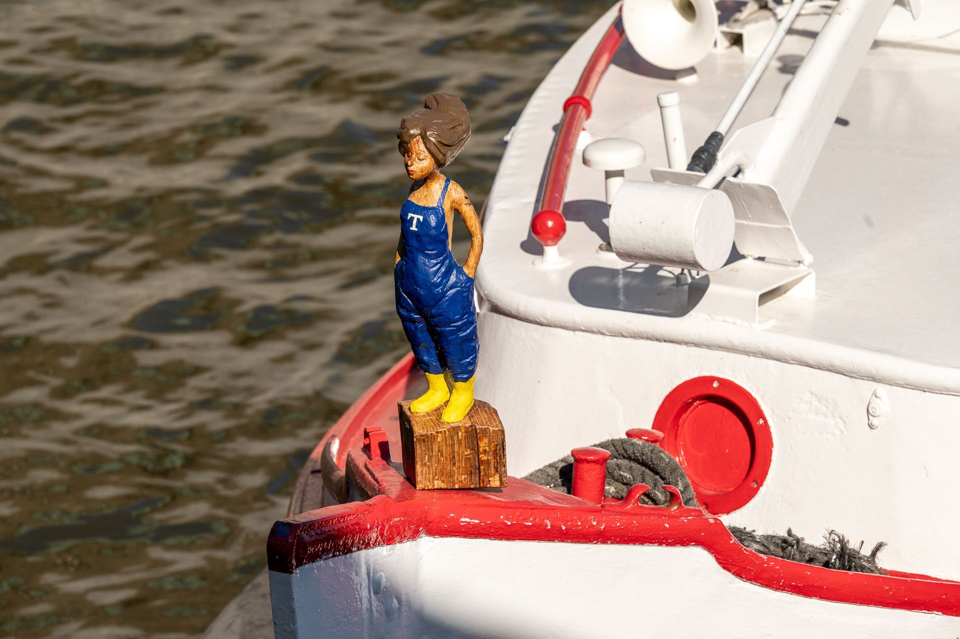 Auf dem Bug eines weißen Bootes steht eine Figur, die eine Person in blauer Latzhose und gelben Stiefeln darstellt.