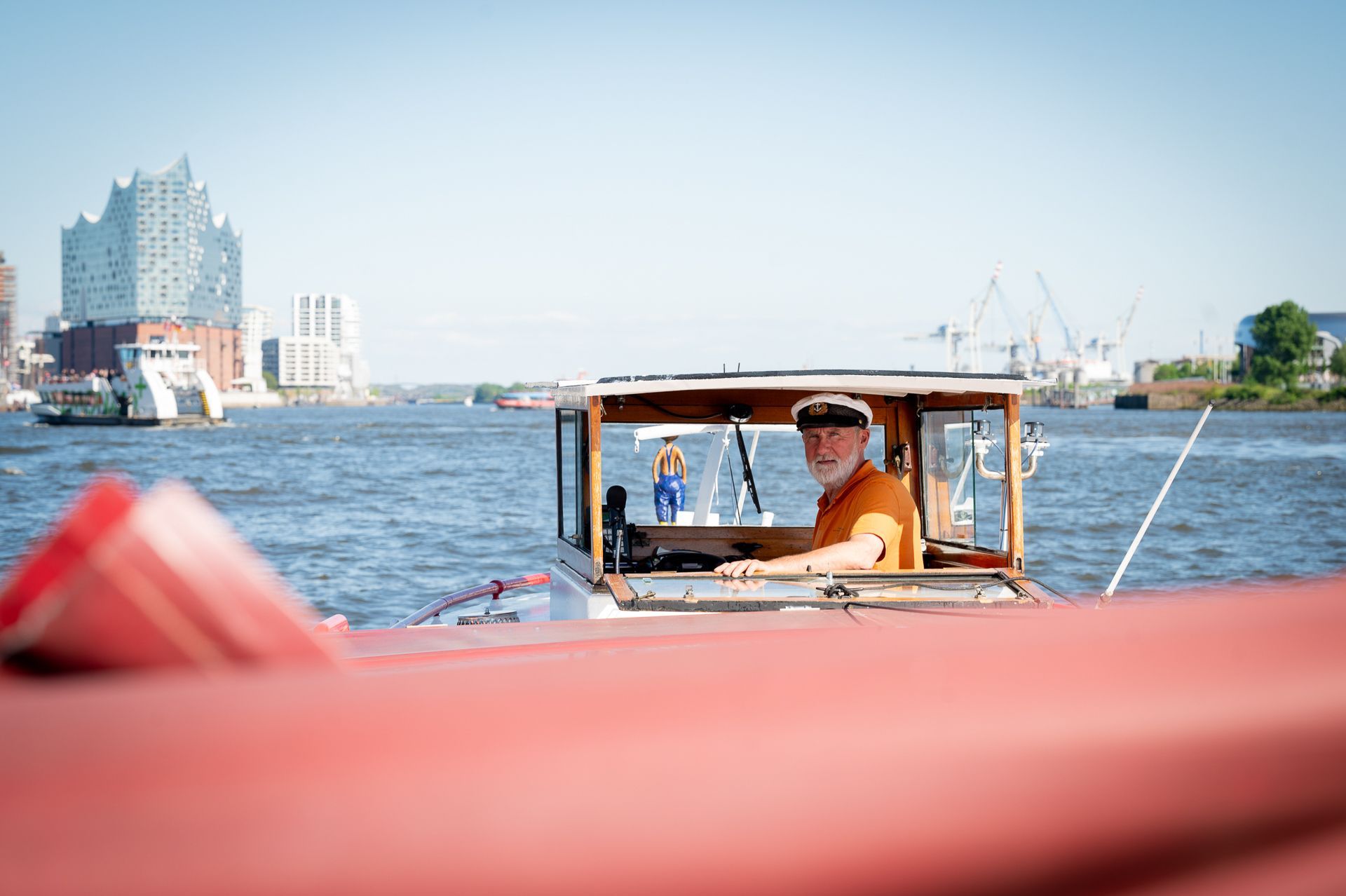 Ein Mann in einem Boot lächelt, während er auf dem Wasser navigiert; im Hintergrund ist eine Stadt mit einem Gebäude zu sehen.