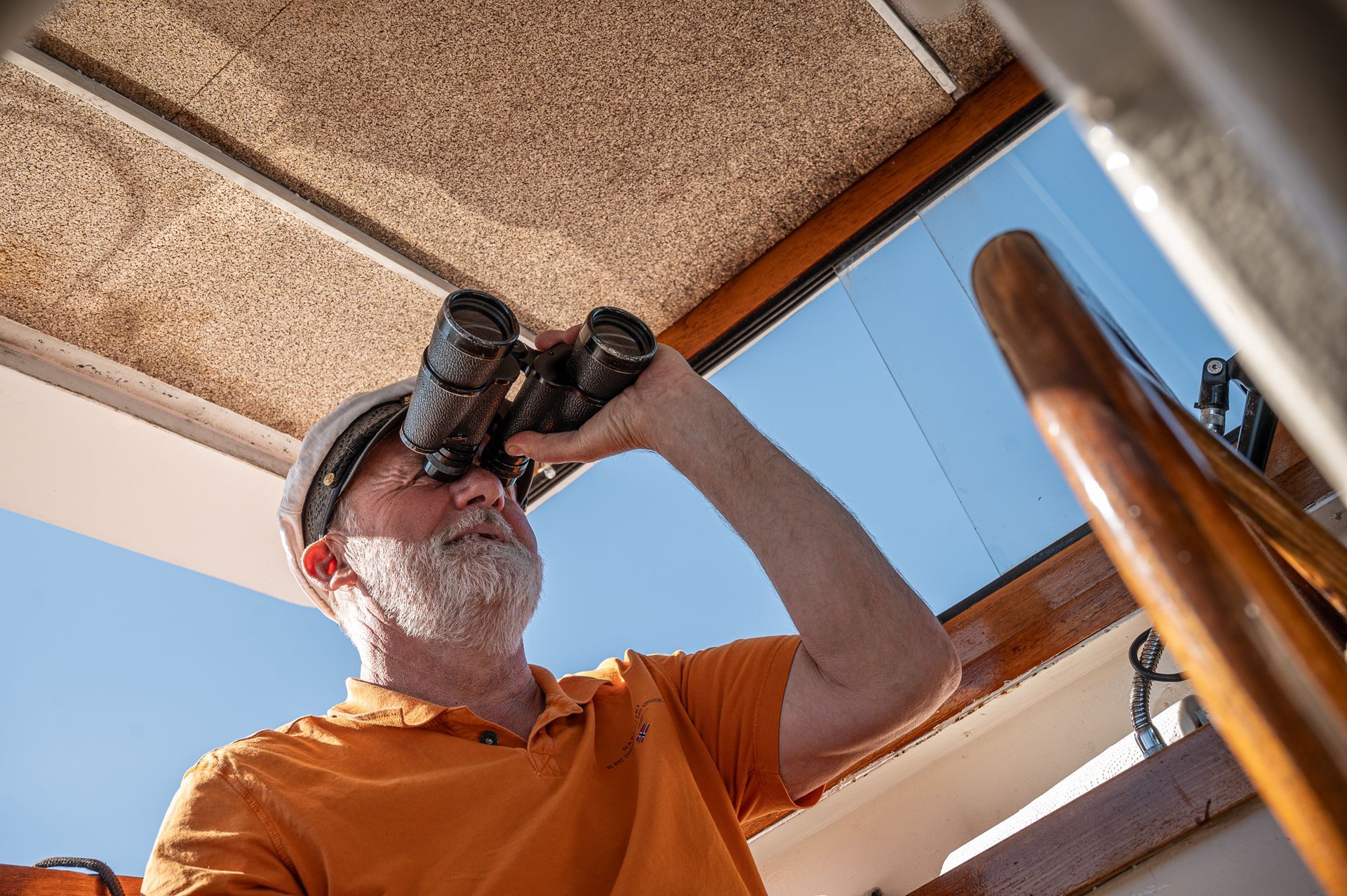 Ein Mann mit Fernglas schaut von einem Boot aus hinaus; er trägt ein orangefarbenes Hemd und einen Schutzhelm.