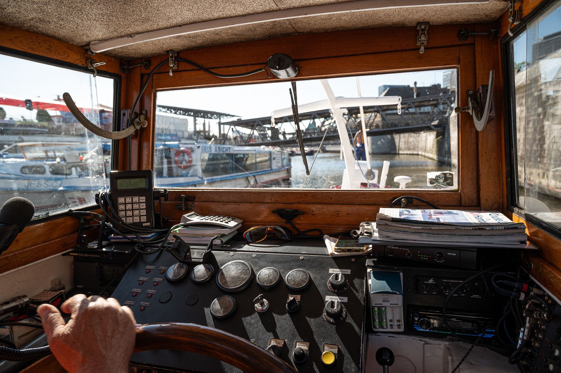 Blick aus der Bootskabine, Steuerrad und Armaturenbrett sind sichtbar. Durch das Fenster erstreckt sich eine sonnige Hafenszene.