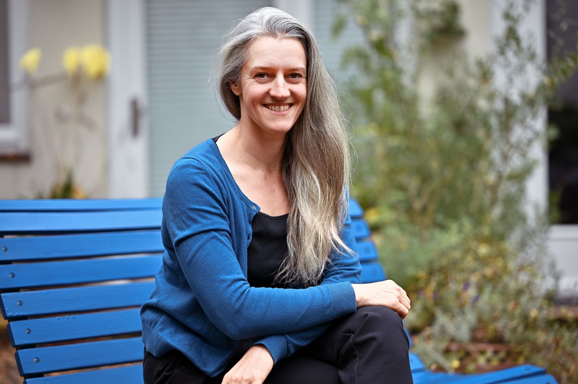 Eine Frau mit langen grauen Haaren lächelt und sitzt draußen auf einer blauen Bank. Sie trägt eine blaue Strickjacke und eine schwarze Hose.