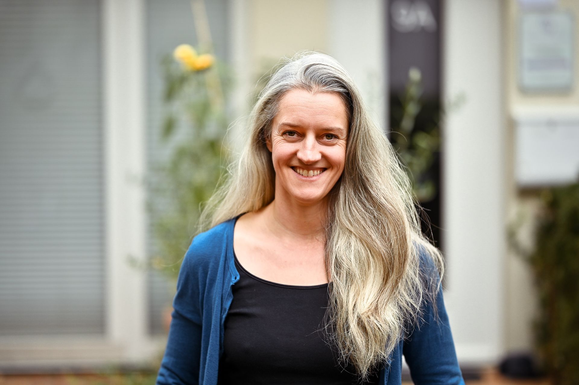 Eine Frau mit langem, ergrauendem Haar lächelt vor einem Gebäude. Sie trägt eine blaue Strickjacke und ein schwarzes Hemd.