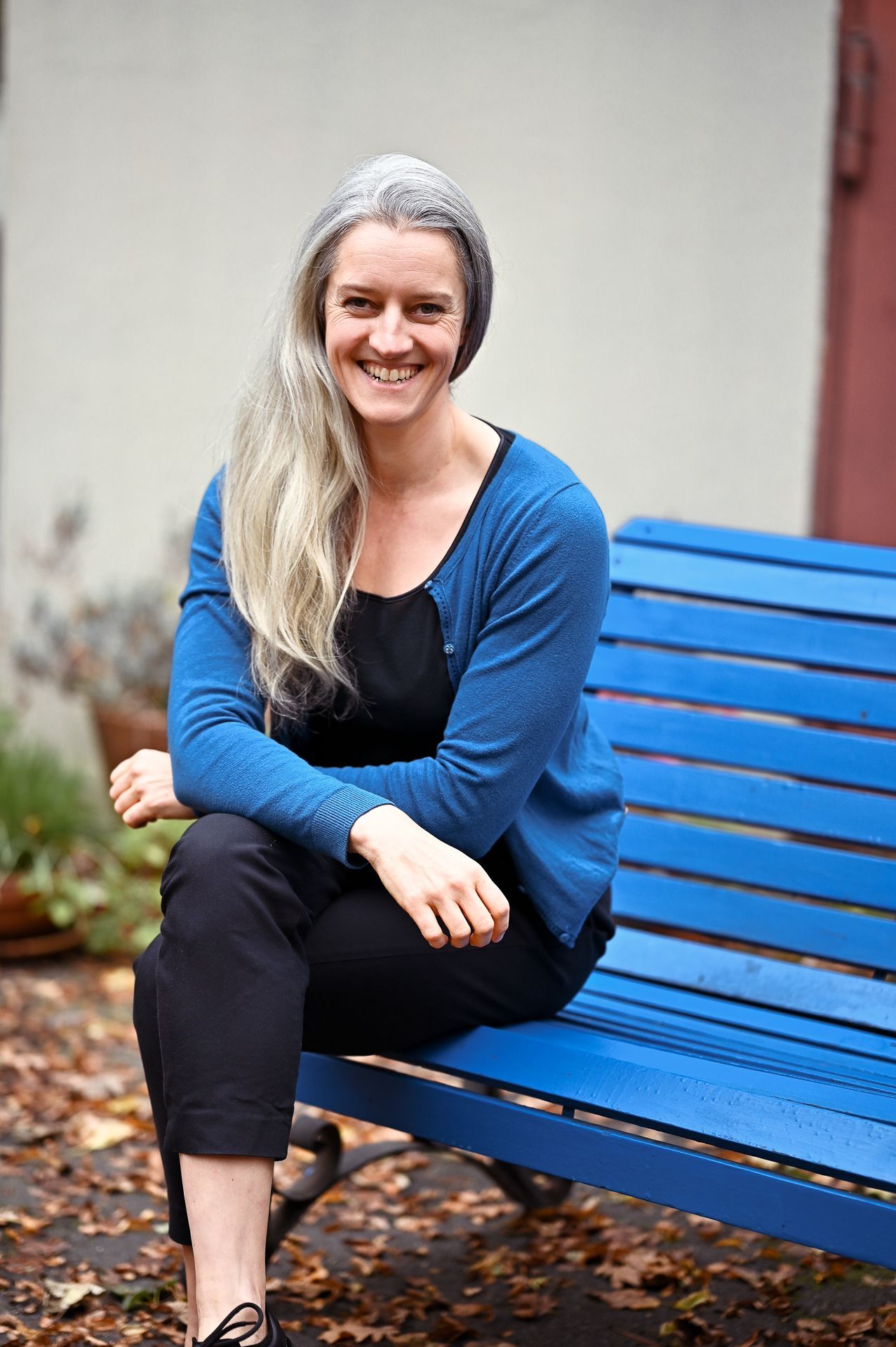 Eine Frau mit langen grauen Haaren lächelt, sie sitzt auf einer blauen Bank und trägt eine blaue Strickjacke und eine schwarze Hose.