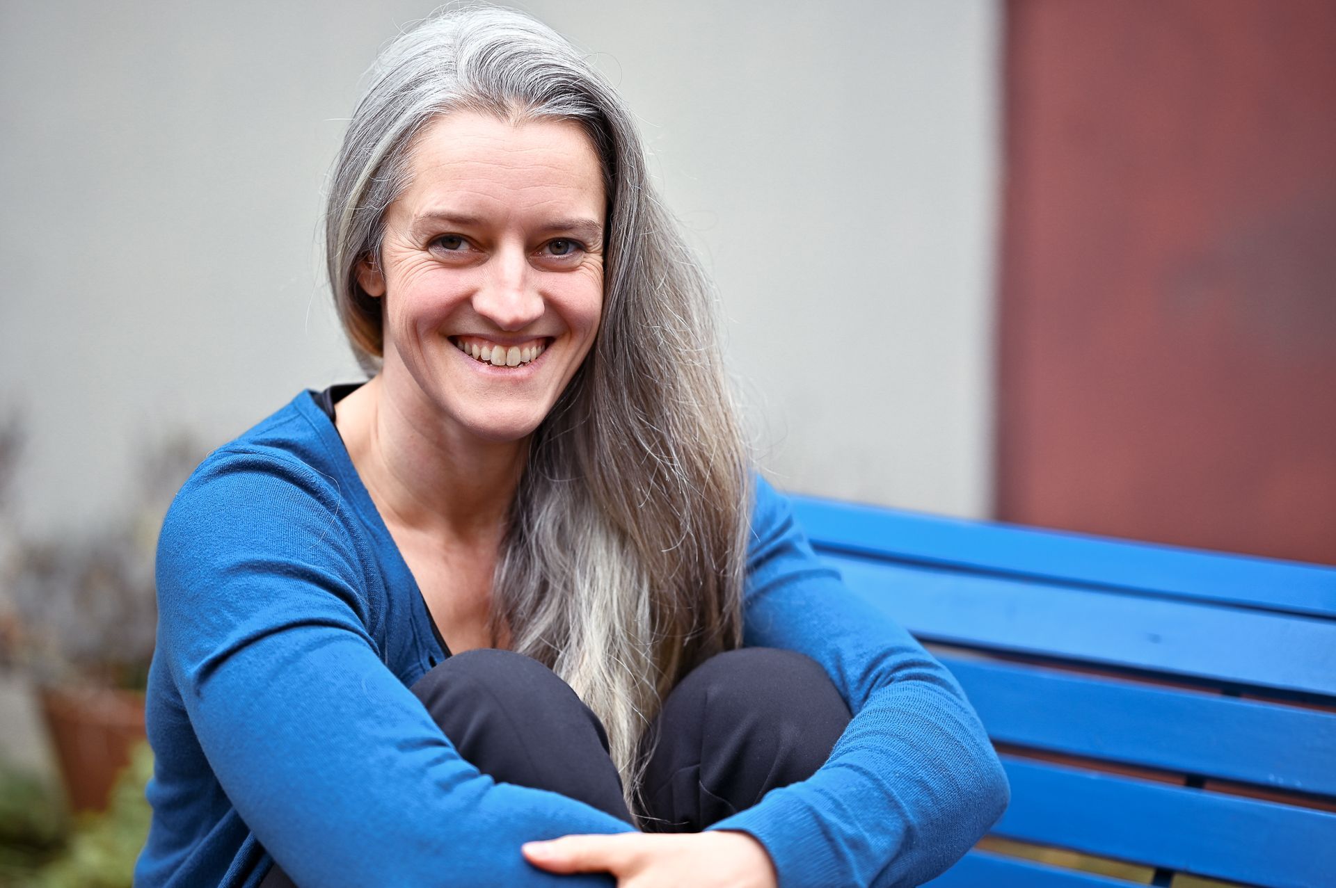 Eine Frau mit langen, grauen Haaren und einem blauen Pullover lächelt, während sie auf einer blauen Bank sitzt.