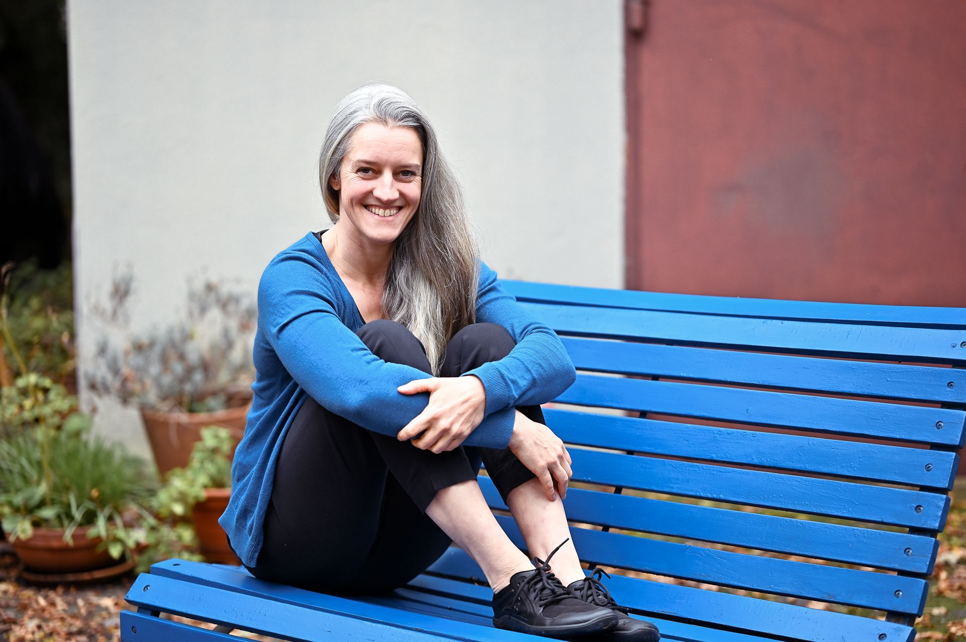 Eine Frau mit grauem Haar lächelt, sitzt auf einer blauen Bank und trägt ein blaues Hemd und eine schwarze Hose.