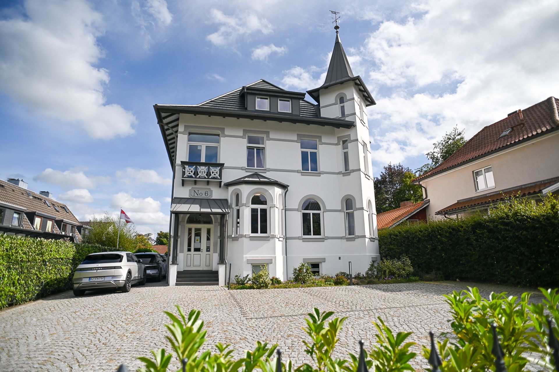 Weißes mehrstöckiges Haus mit Turm und Bogenfenstern, gepflasterte Auffahrt, blauer Himmel.
