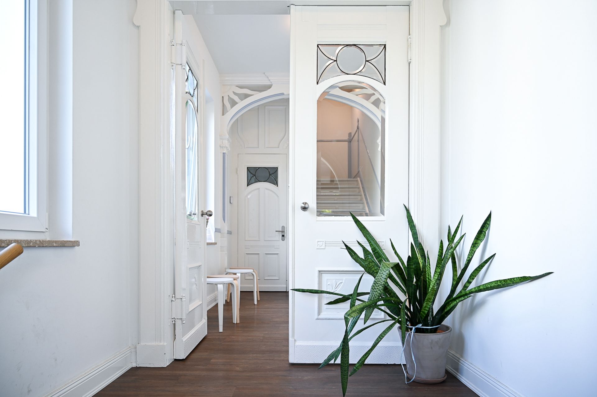 Ein strahlend weißer Flur mit verzierten Türen, zwei Stühlen und einer Topfpflanze führt zur Treppe.