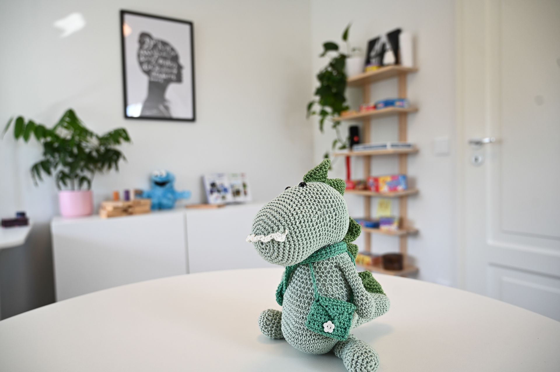 Gehäkeltes Dinosaurier-Spielzeug auf einem weißen Tisch in einem Zimmer mit einem weißen Schrank, Pflanzen und Kunstwerken.