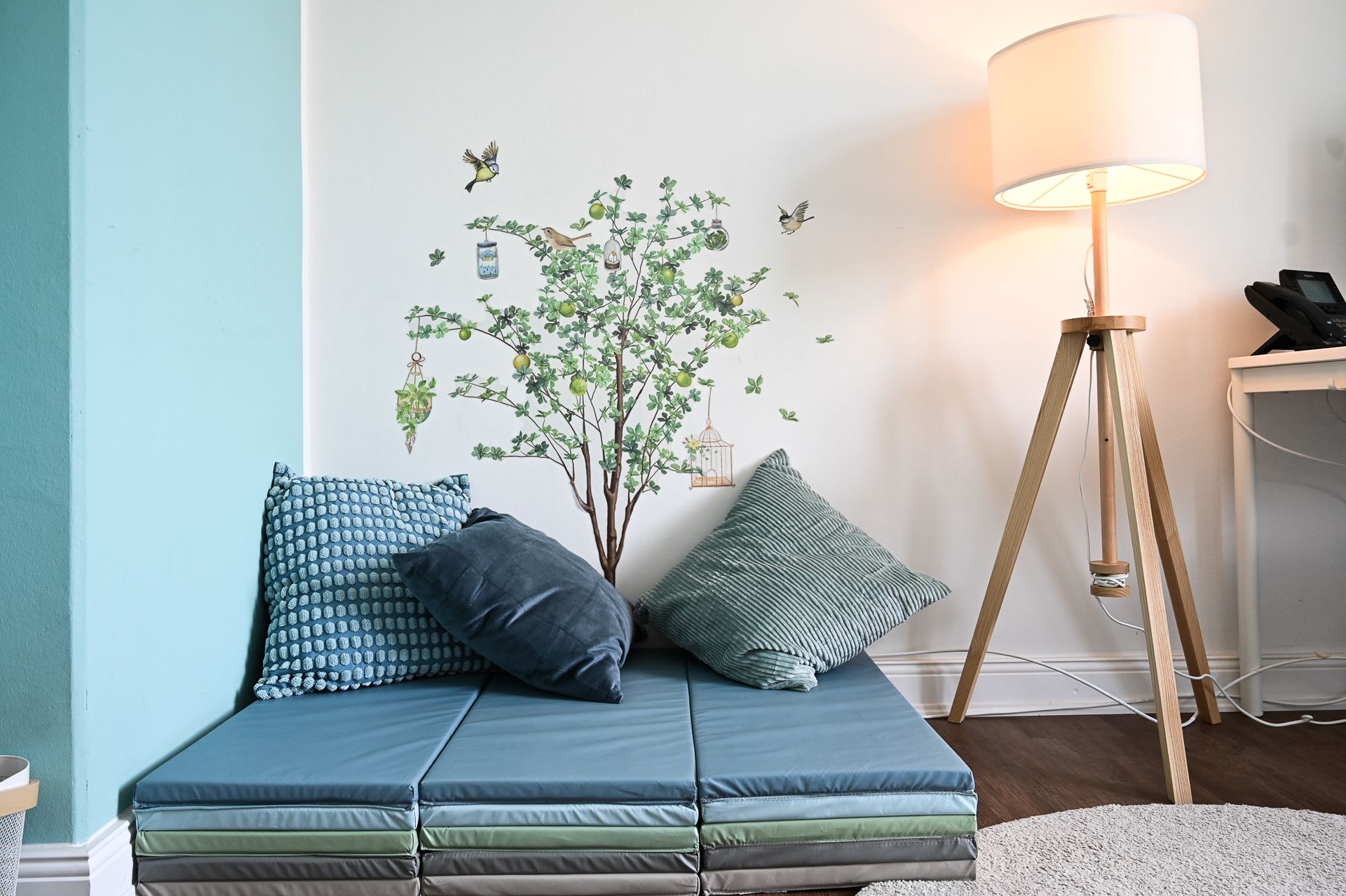 Ein blaues, gepolstertes Tagesbett mit Kissen vor einem Baum-Aufkleber an einer weiß-hellblauen Wand, neben einer Stehlampe.