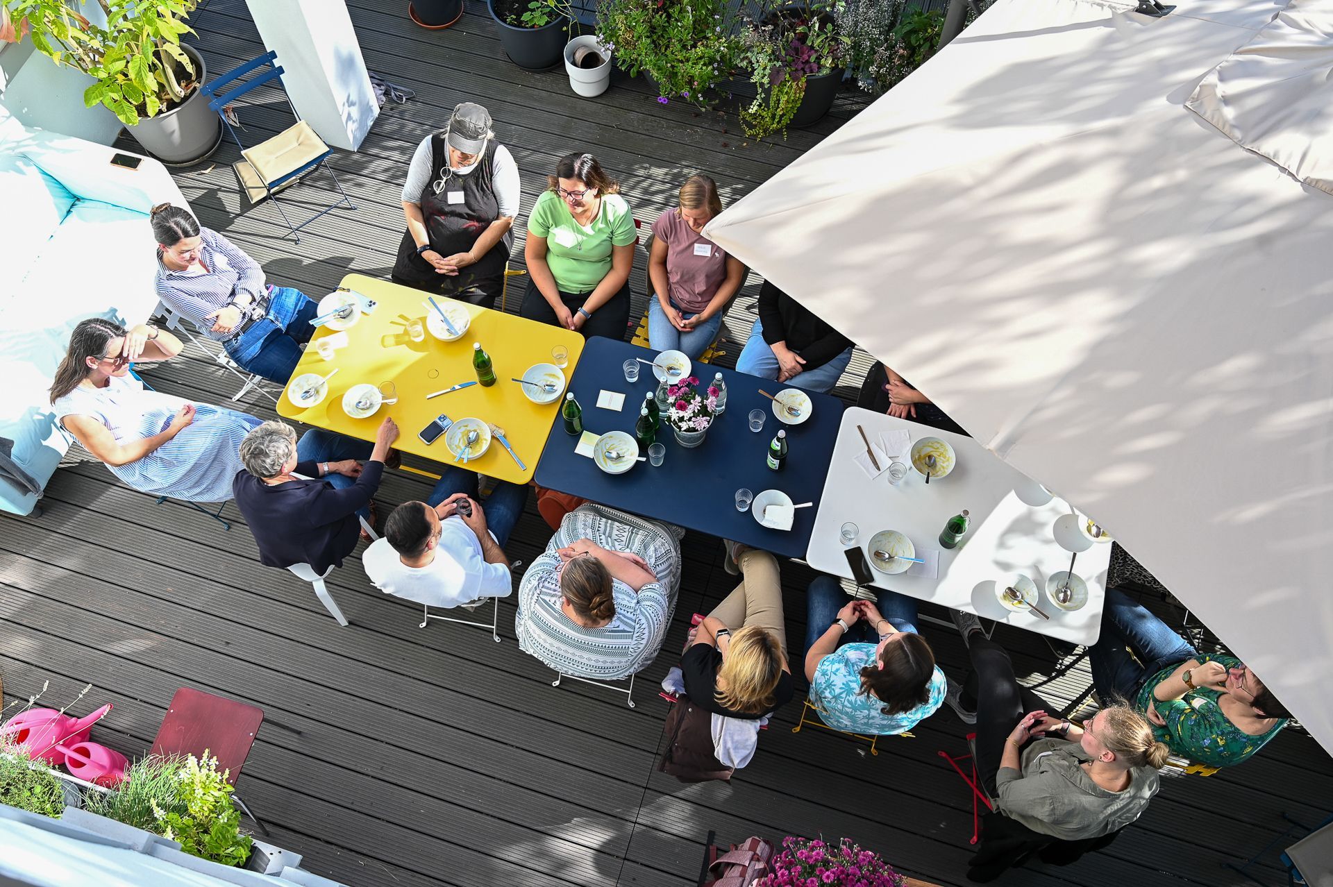 Eine Gruppe von Menschen sitzt an Tischen auf einer Terrasse, mit Essen und Getränken. Aufnahme von oben.