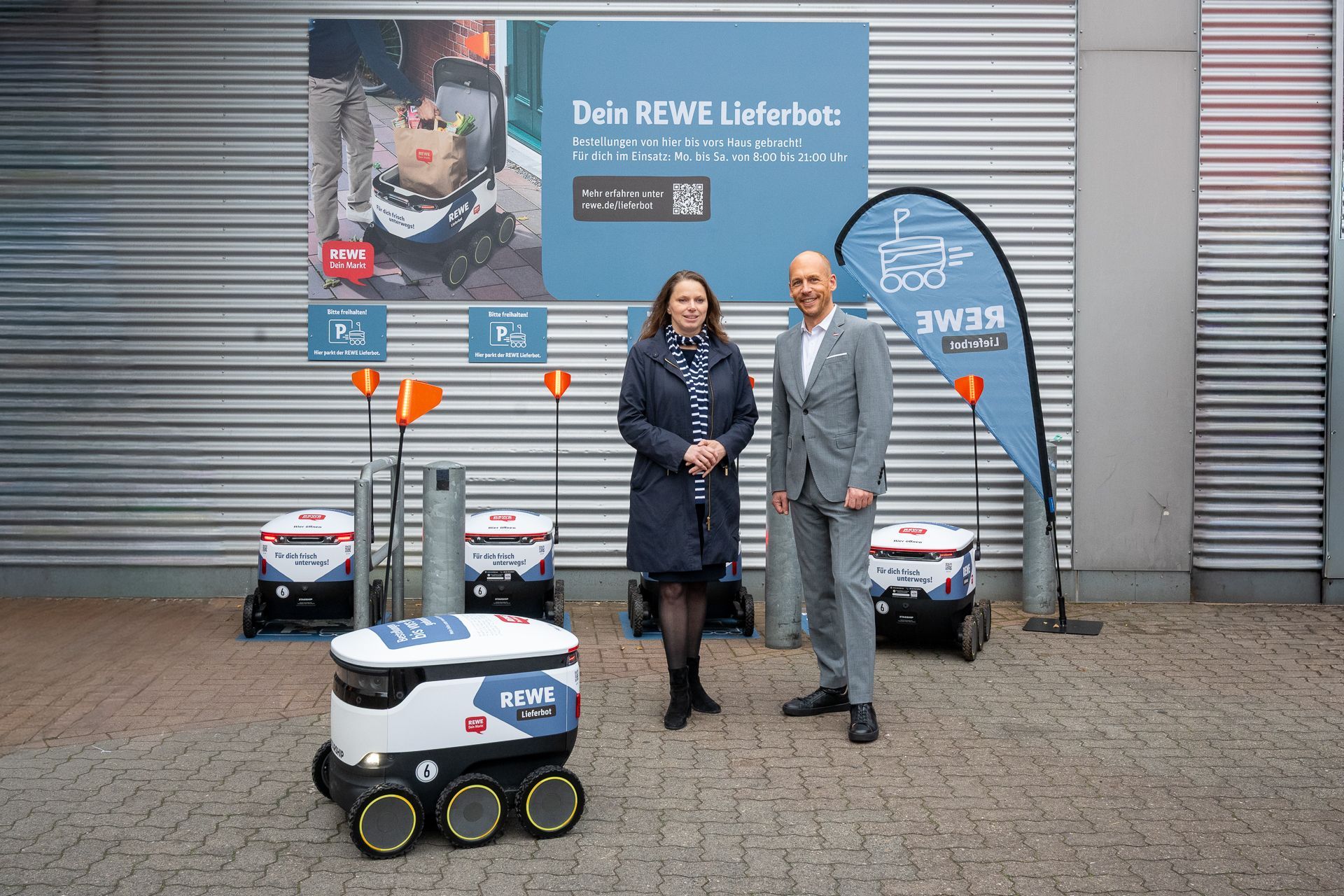 Zwei Personen stehen neben Lieferrobotern vor einem Gebäude mit Hinweisschildern.