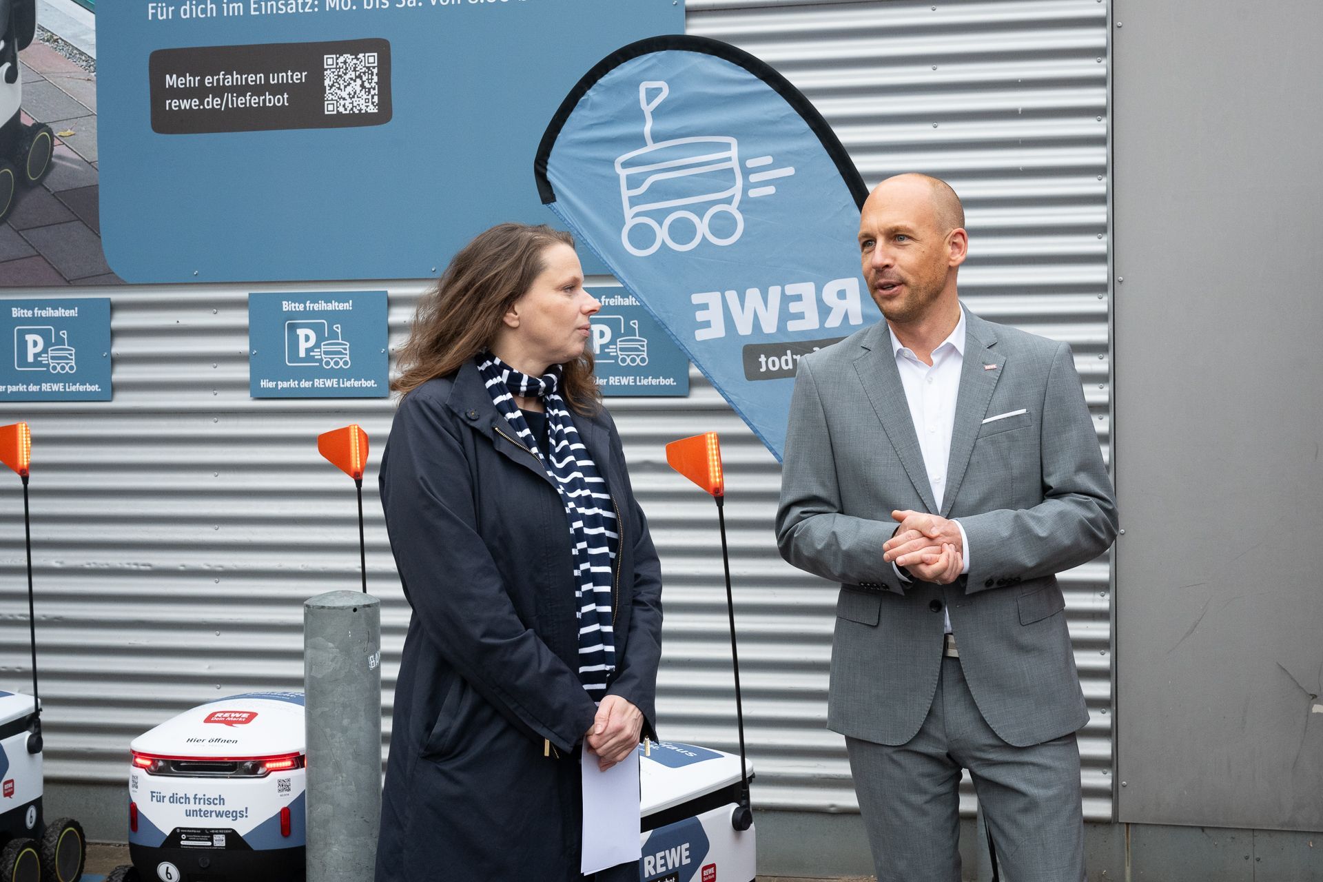 Zwei Personen stehen neben Lieferrobotern. Ein Mann im Anzug spricht, während eine Frau in einer Jacke zusieht.