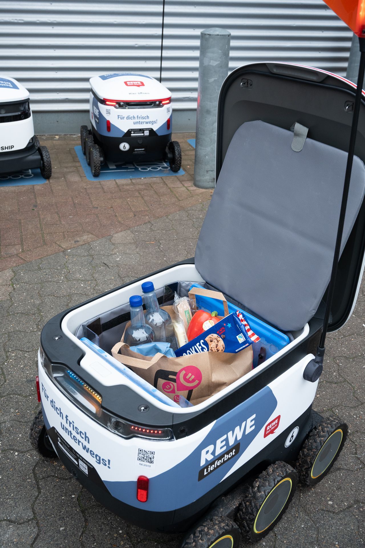 Lieferroboter für Lebensmittel mit geöffnetem Deckel, der die Lebensmittel freigibt. REWE-Logo sichtbar. Im Freien auf dem Gehweg.
