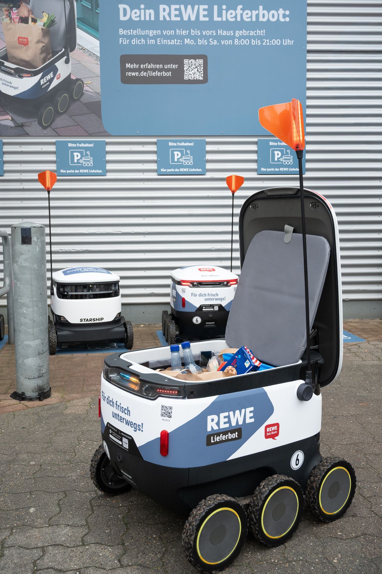 REWE-Lieferroboter mit offenen Laderäumen parken vor einem Geschäft mit Beschilderung.