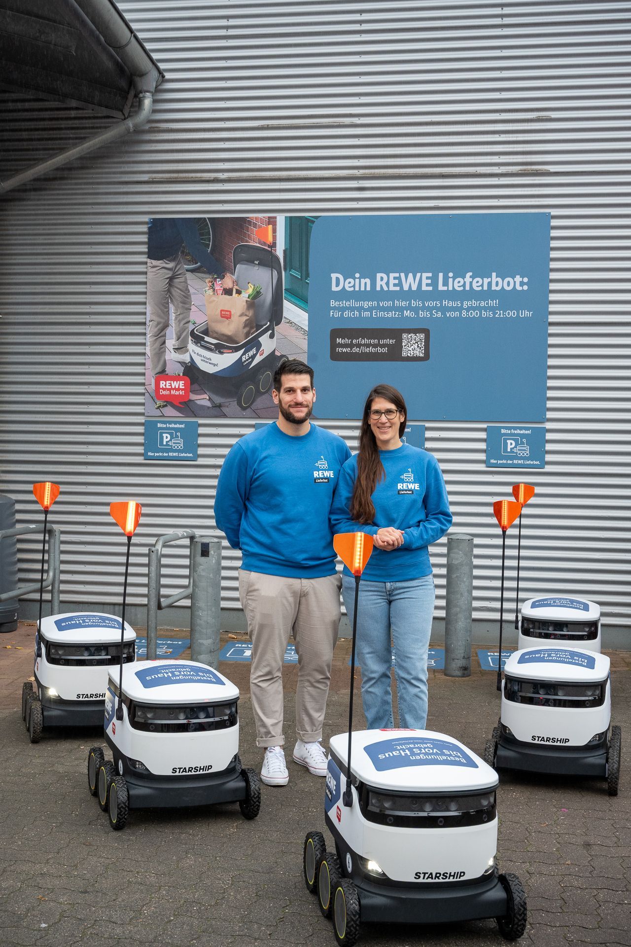 Zwei Personen in blauen Hemden stehen mit Lieferrobotern vor einem REWE-Schild.