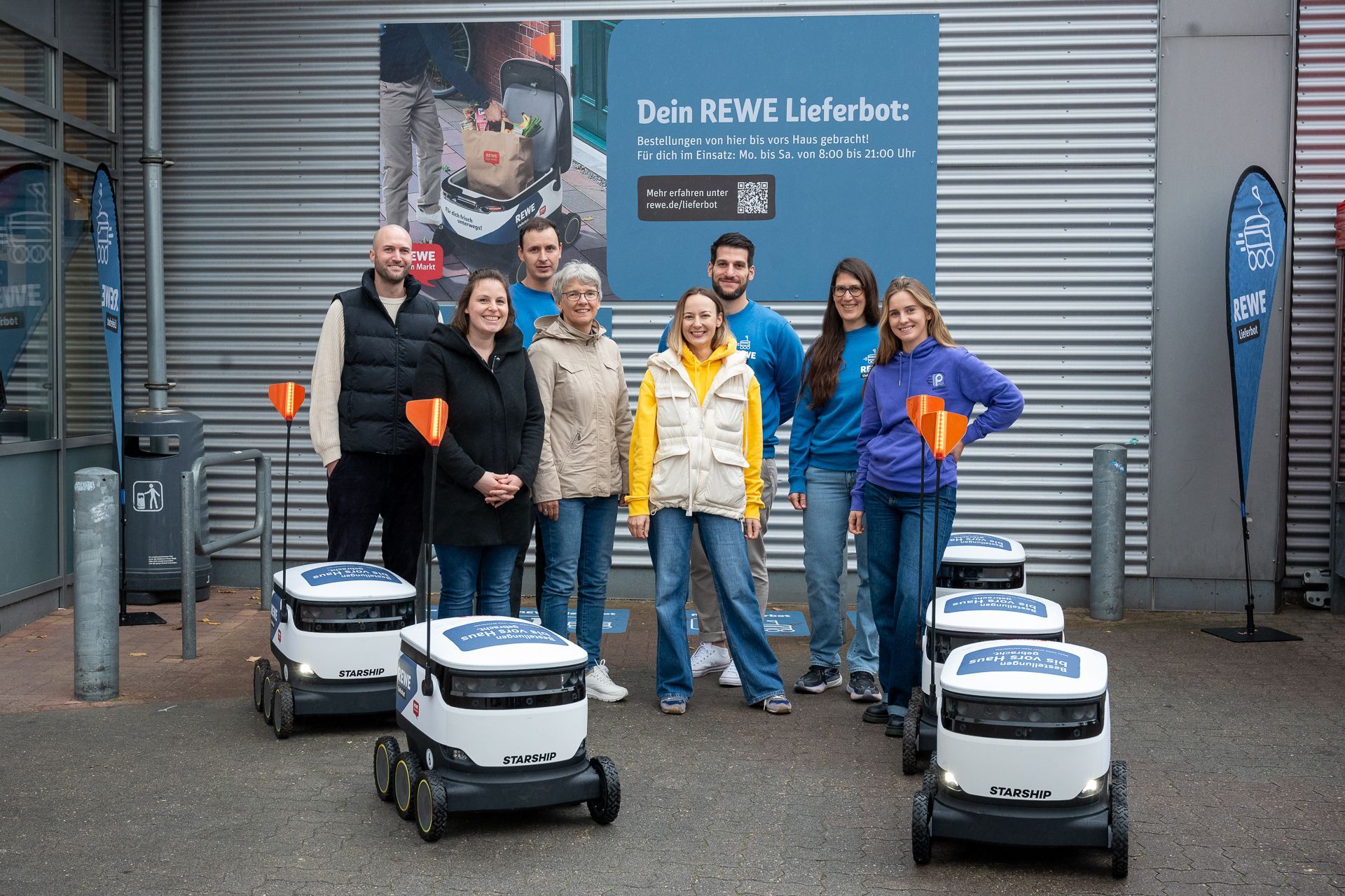 Menschen posieren mit Lieferrobotern vor einem REWE-Supermarkt.