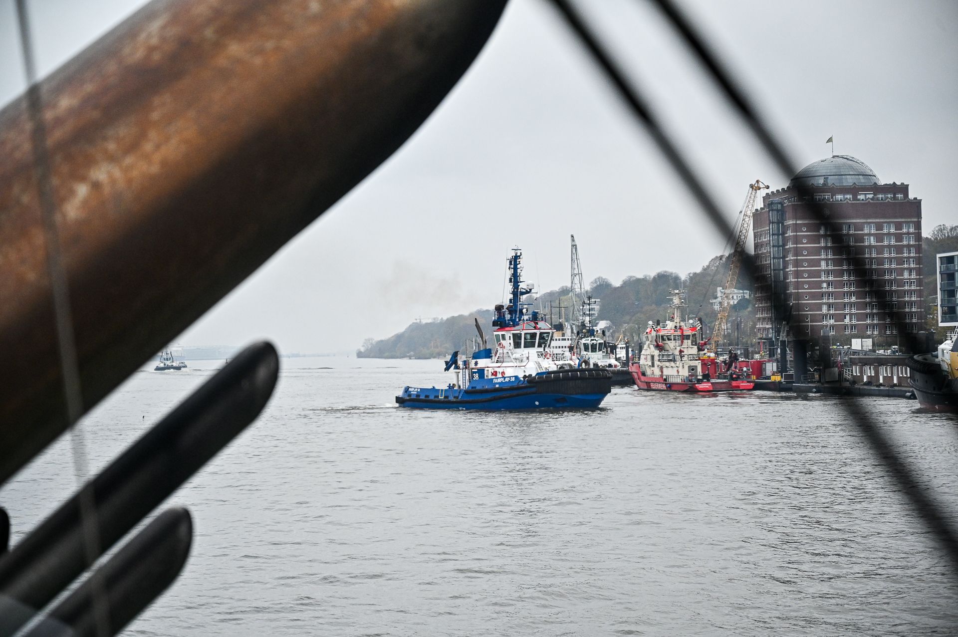Ein blau-weißer Schlepper auf dem Wasser, eingerahmt von der Takelage eines Schiffes, im Hintergrund Gebäude und Bäume.