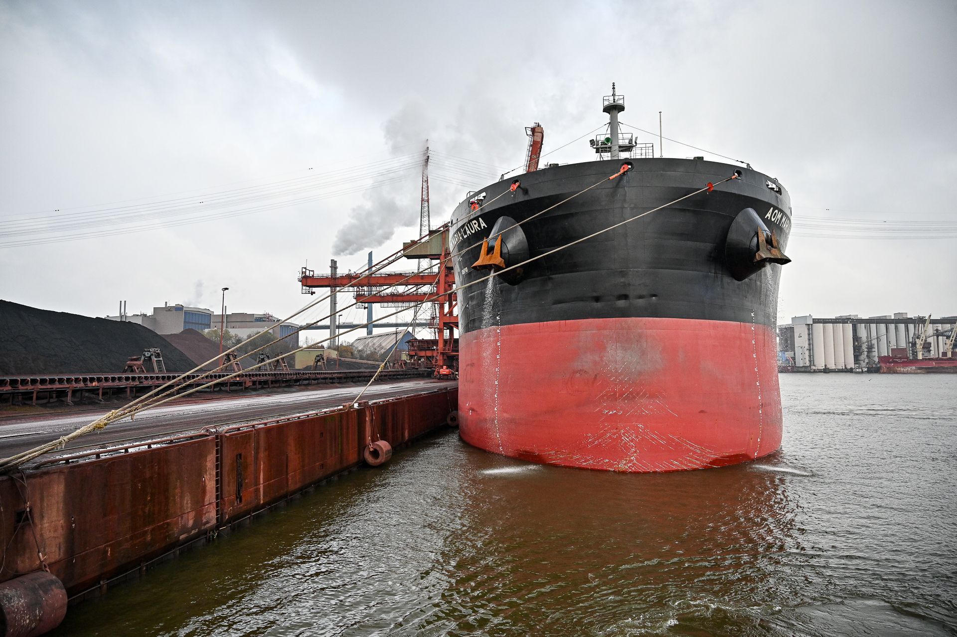 Ein großes, schwarz-rotes Frachtschiff liegt an einer Verladeanlage vor Anker. Im Hintergrund sind Kohle- und Industriegebäude zu sehen.