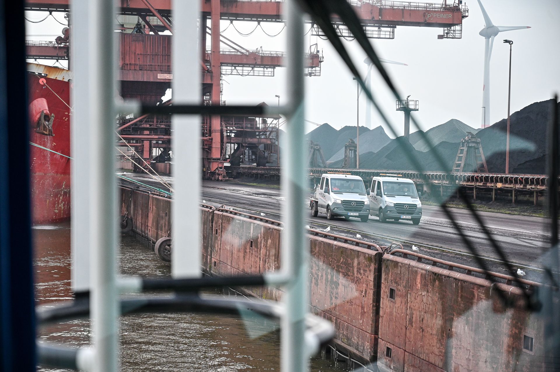 Blick aus der Fahrerkabine eines Fahrzeugs: zwei Lieferwagen auf einem Pier neben einem Frachtschiff und Kohlehaufen.