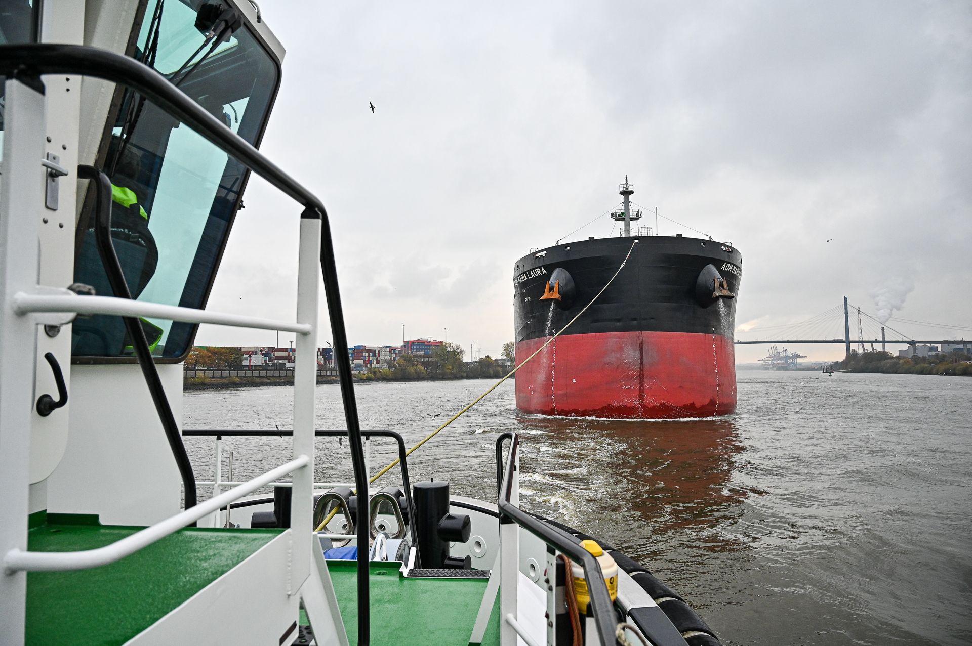 Schlepper im Vordergrund, im Wasser ein großer schwarz-roter Tanker an einem bewölkten Tag.
