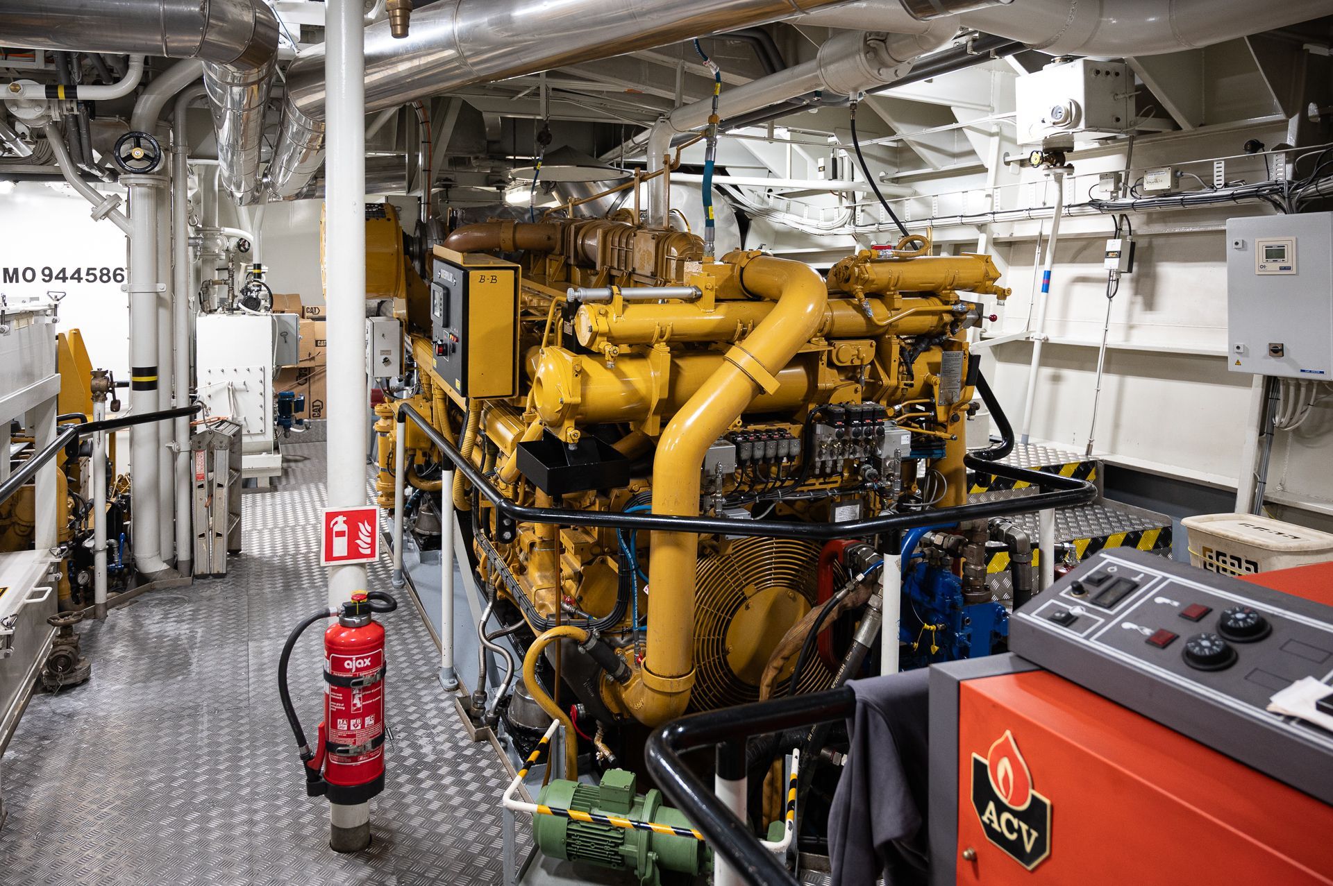 Gelber Schiffsmotor im Maschinenraum eines Schiffes, mit Rohren, Feuerlöscher und Bedienfeld.