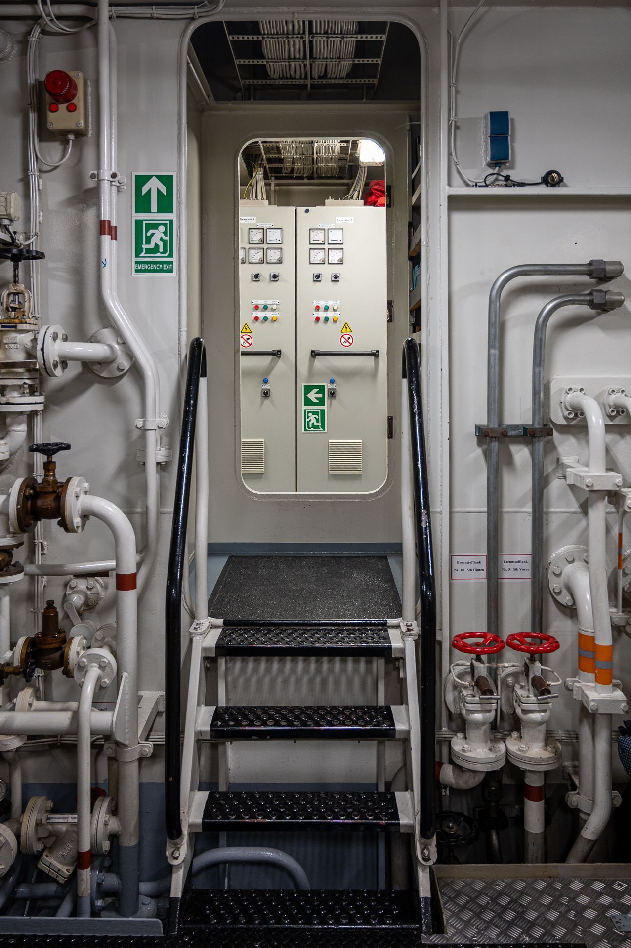 Türöffnung mit Treppe auf einem Schiff, die zu einem Schaltschrankraum führt. Weiße Rohre und Ventile.