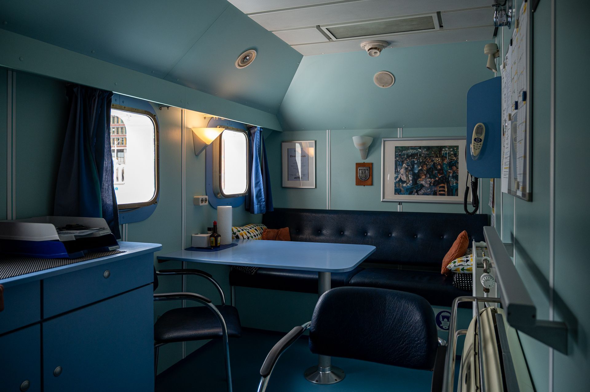 Interieur eines türkisfarbenen Zimmers auf einem Boot oder in einem Zug, mit Tisch, Stühlen, Sofa und kleinen gerahmten Bildern.