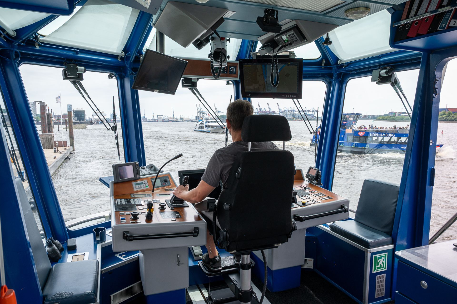 Ein Mann bedient das Bedienfeld eines Bootes; er sitzt auf einem Stuhl; draußen vor dem Fenster sind Wasser und andere Boote zu sehen.