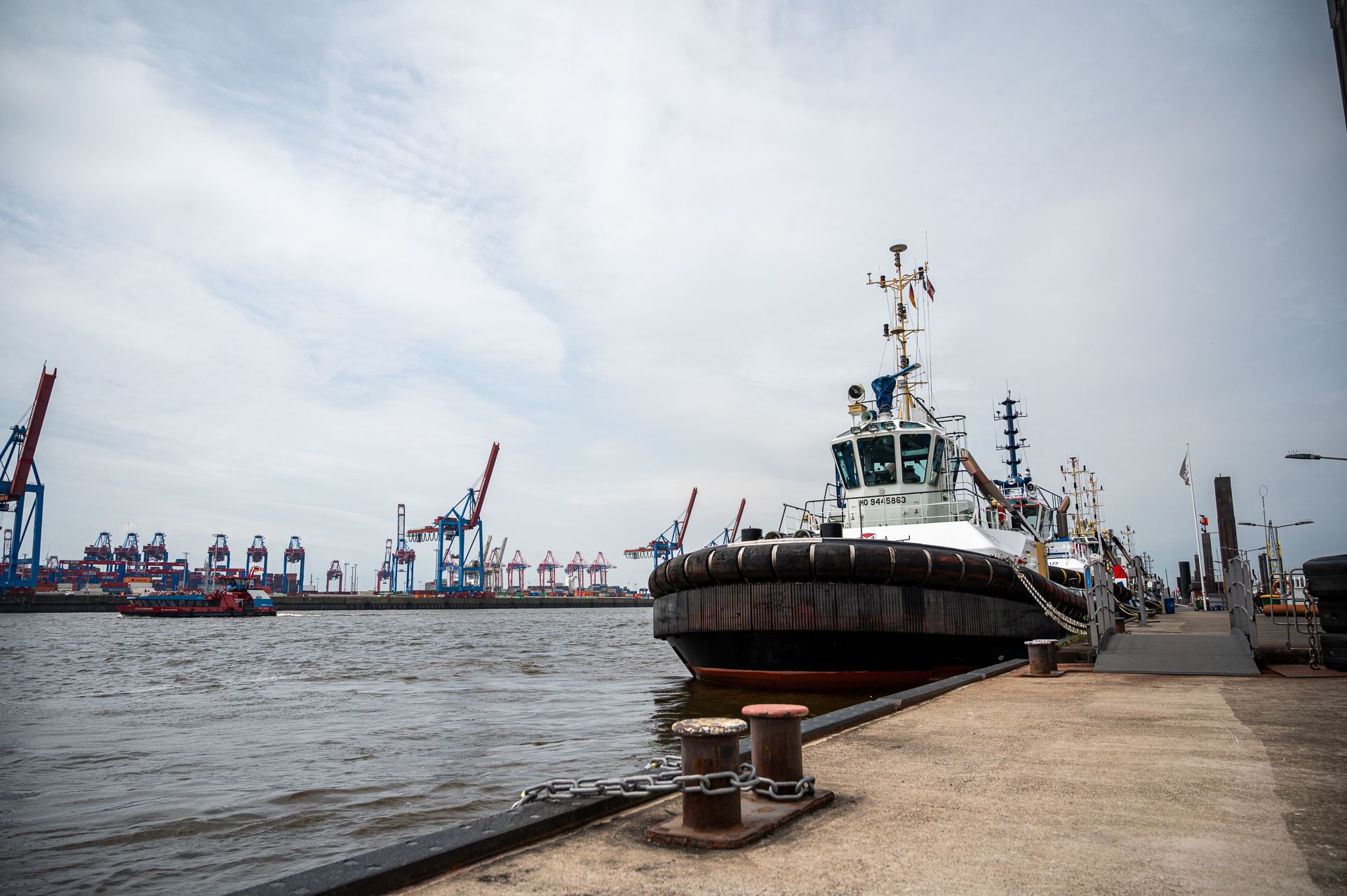 Ein Schlepper liegt im Hafen vor Anker, im Hintergrund sind Industriekräne unter einem bewölkten Himmel zu sehen.
