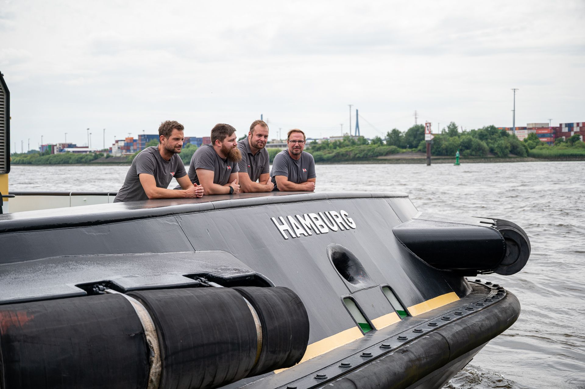 Vier Männer auf einem schwarzen Schlepper blicken in die Kamera. Auf dem Boot steht „Hamburg“. Wasser- und Industrieumgebung.