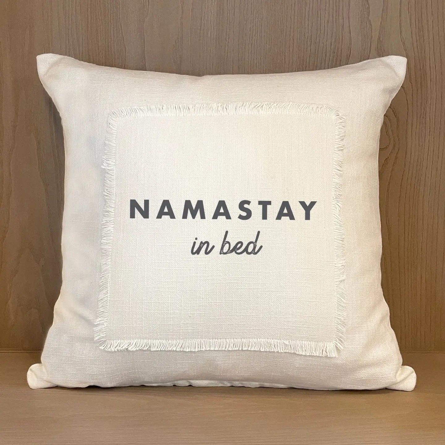 Cream pillow with “NAMASTAY in bed” text on a wooden shelf.