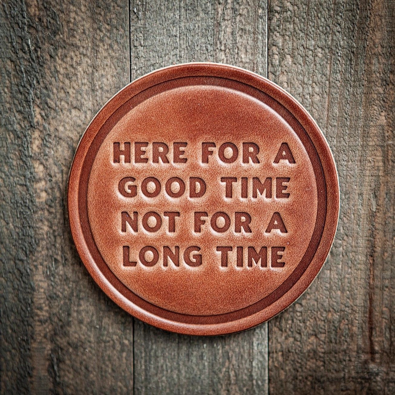 Round rust-colored coaster with text: “Here for a good time, not for a long time” on weathered wood.
