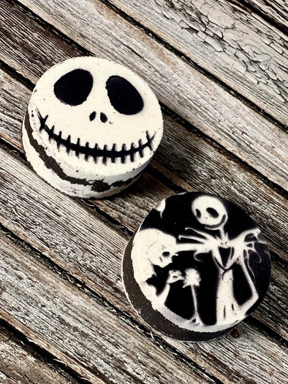 Two black-and-white round pins on weathered wood, one with a Jack Skellington face and one with a skeleton scene