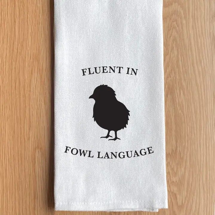 White towel with a black chicken silhouette and the text “FLUENT IN FOWL LANGUAGE” on a wood background