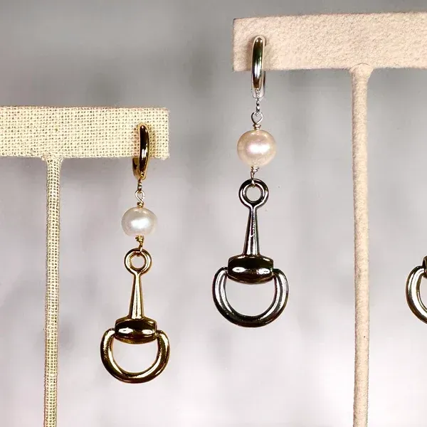 Two dangling pearl-and-metal earrings displayed on beige stands.